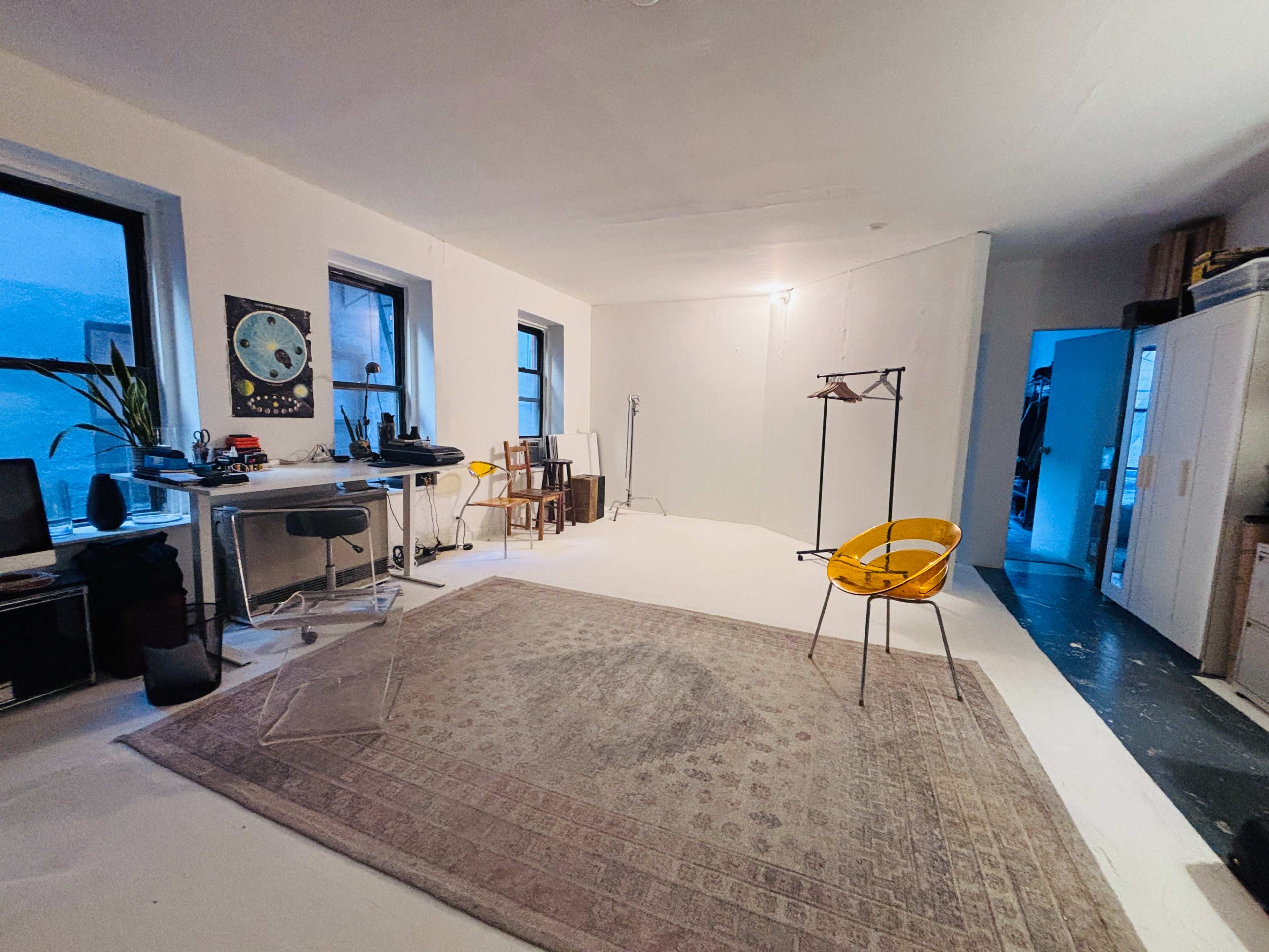 The image shows a spacious, empty room with large windows, a circular rug, a yellow chair, a desk with equipment, and a closet in the corner.