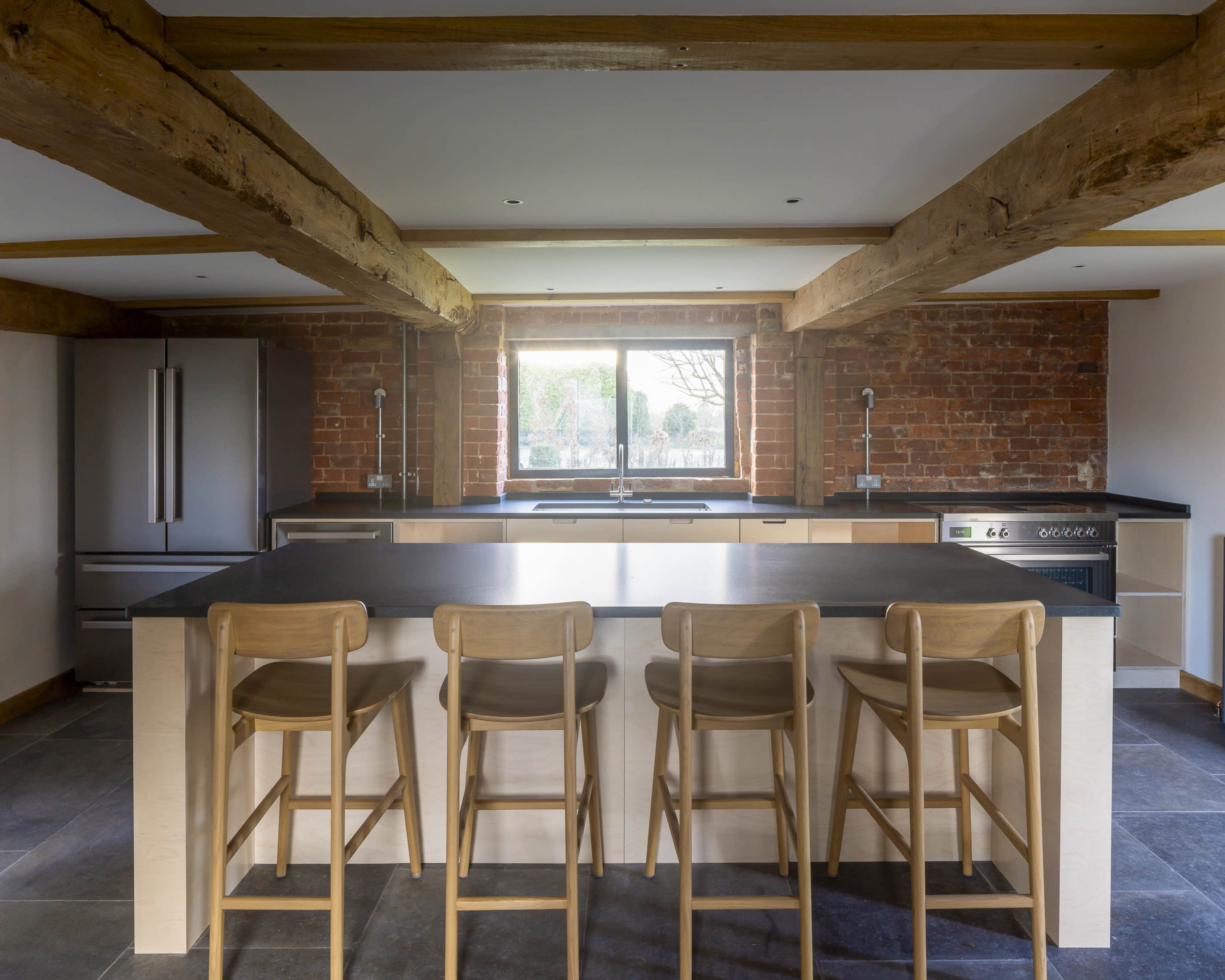 The image depicts a modern kitchen with a central island and four wooden bar stools, featuring exposed brick walls and wooden beams.