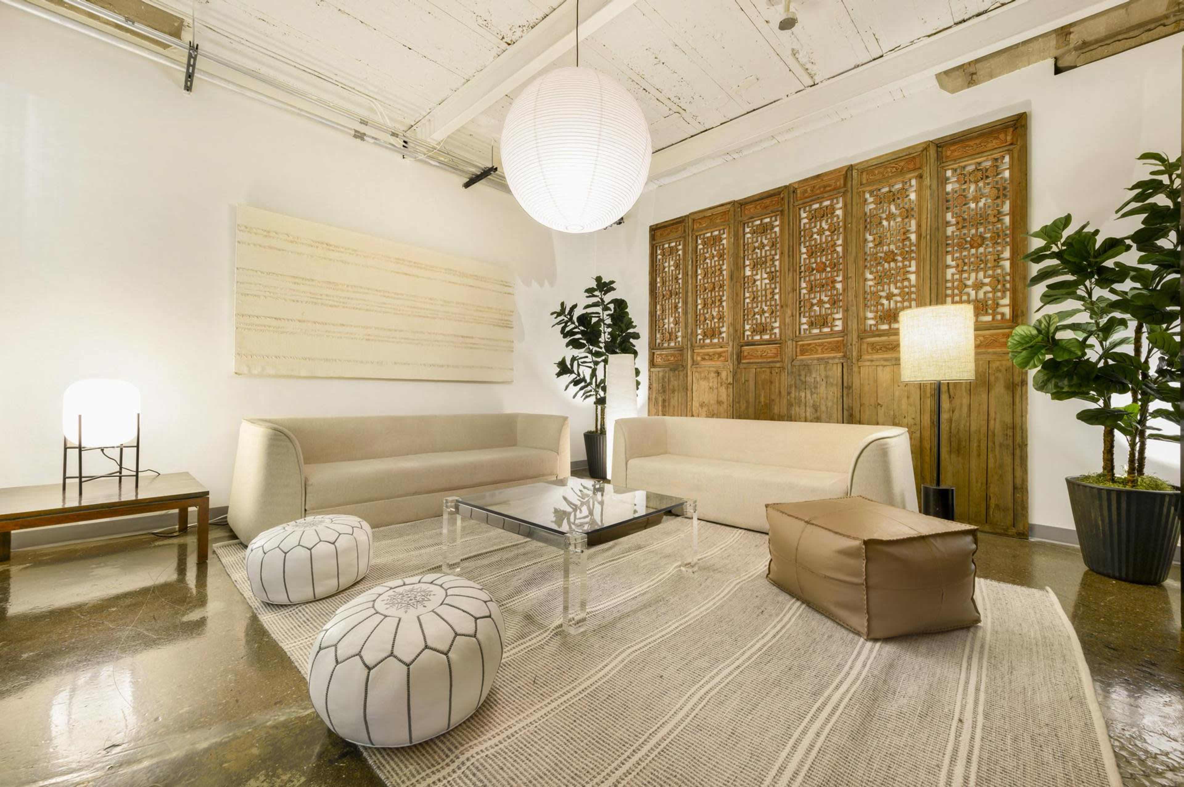 The image shows a modern living room with two sofas, a glass coffee table, decorative poufs, and plants, all set against a wooden screen and minimalistic decor.