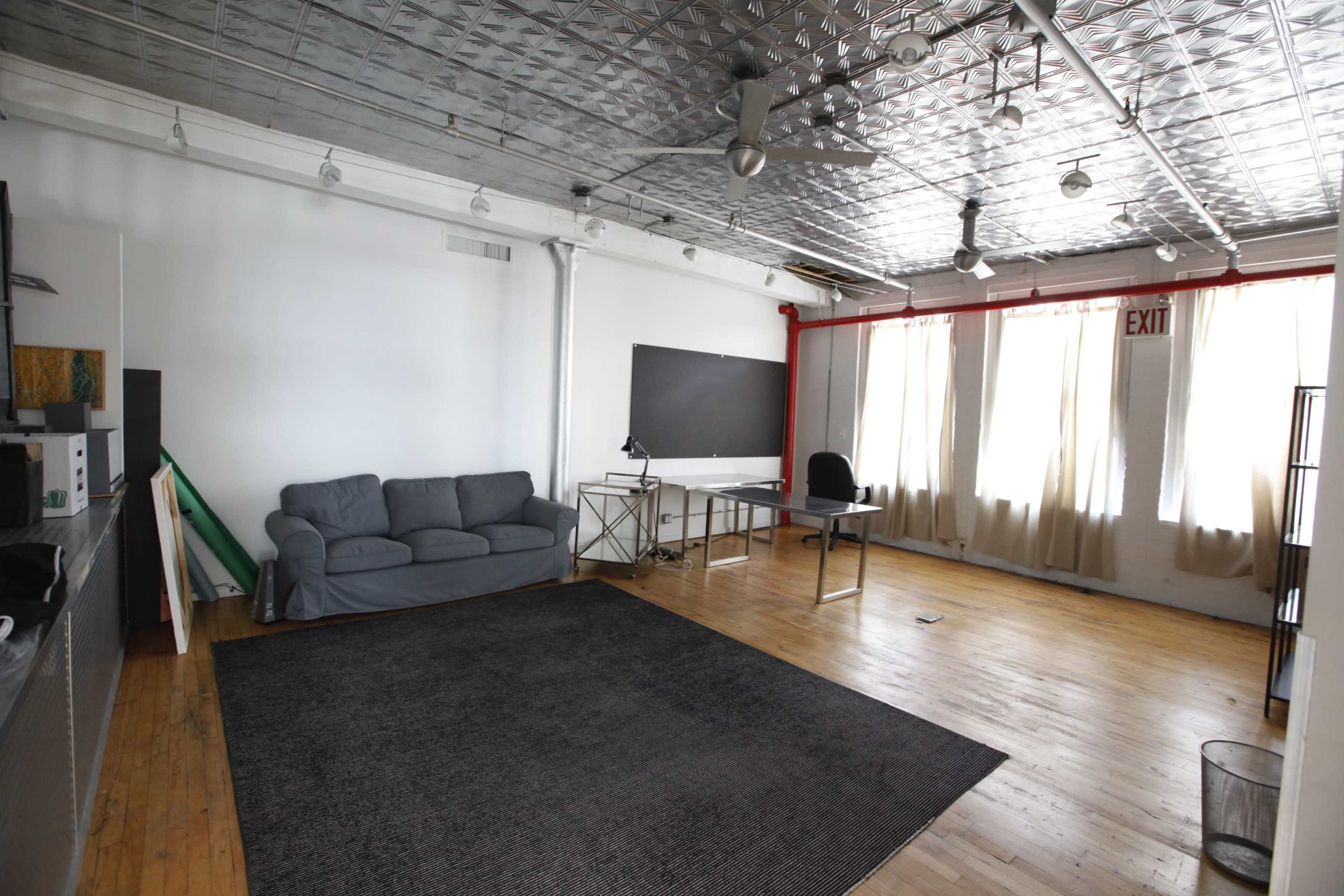 A spacious, empty room featuring a gray sofa, a desk and chair, large windows, and a metal ceiling.