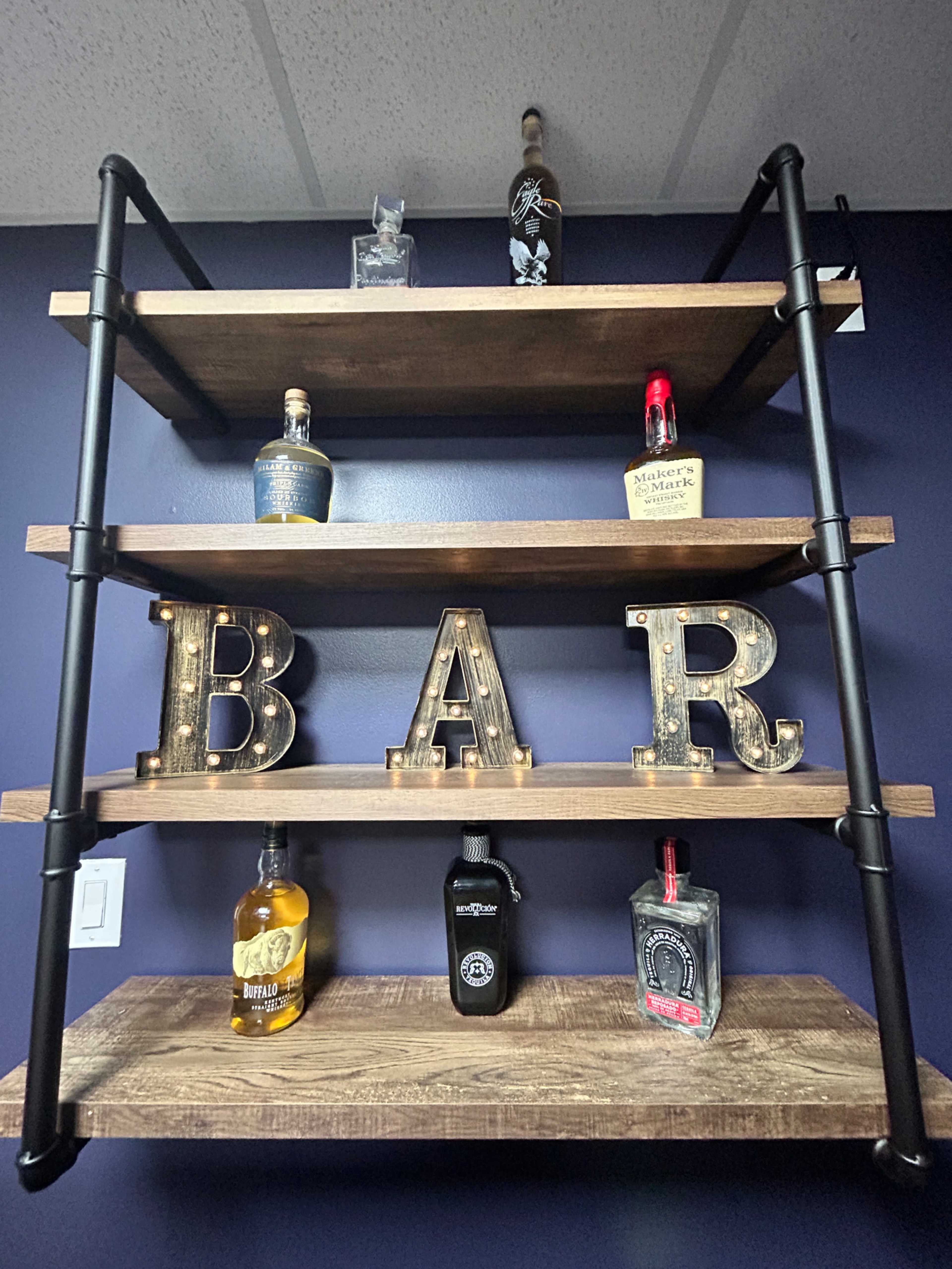 The image features a three-tiered wooden shelf with a dark wall background, displaying various alcohol bottles and illuminated letters spelling "BAR."