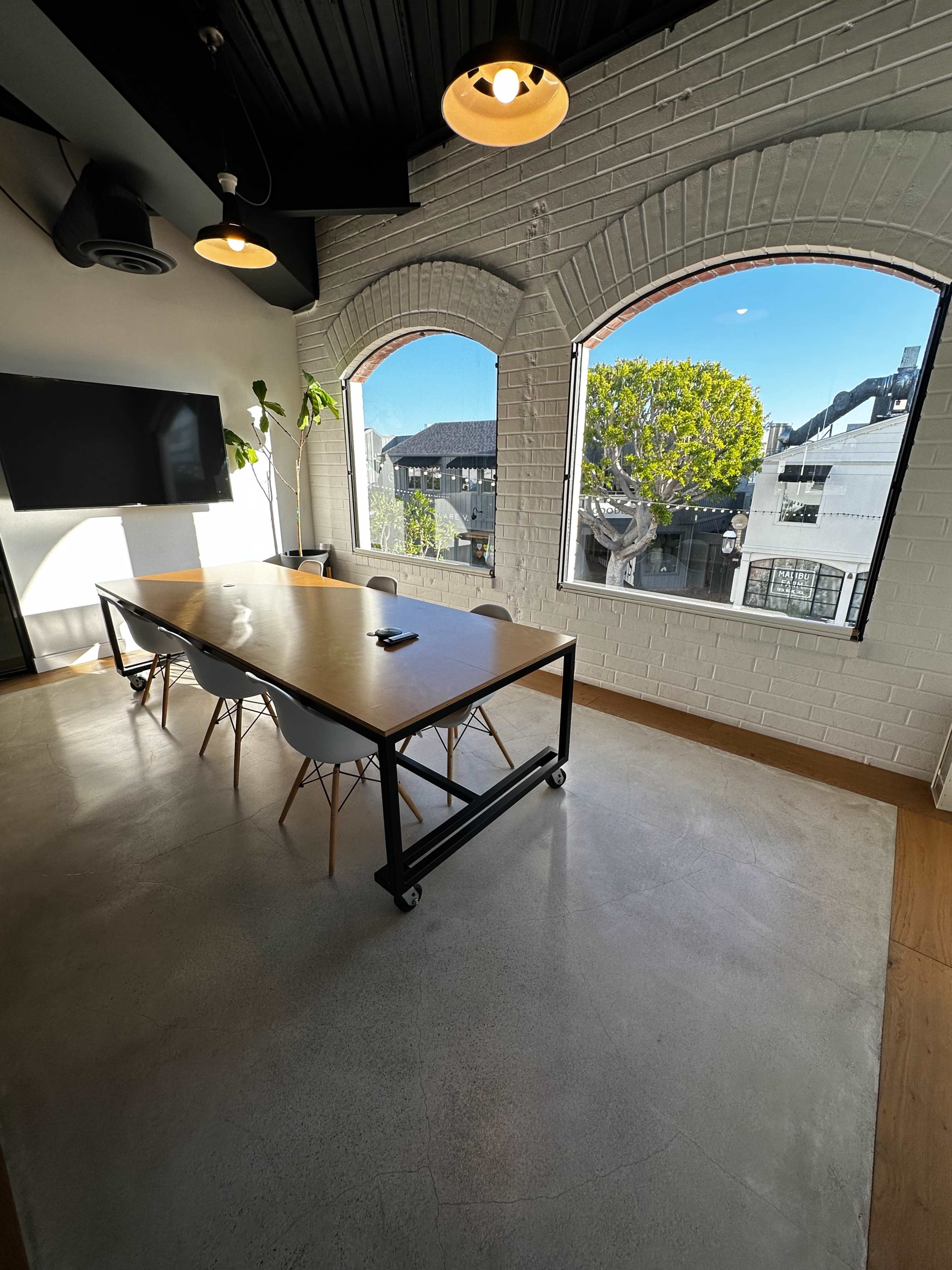 A modern conference room features a large wooden table surrounded by chairs, with large arched windows offering a view of an outdoor area.