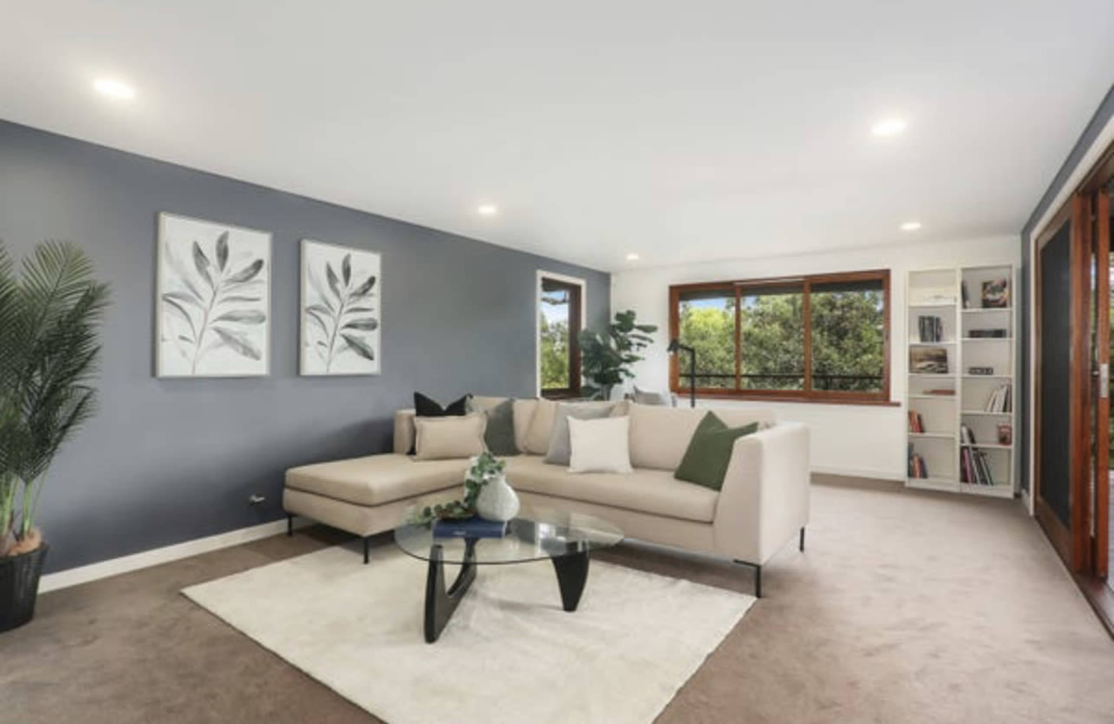 A modern living room features a light-colored sectional sofa, a glass coffee table, and large windows with views of greenery.
