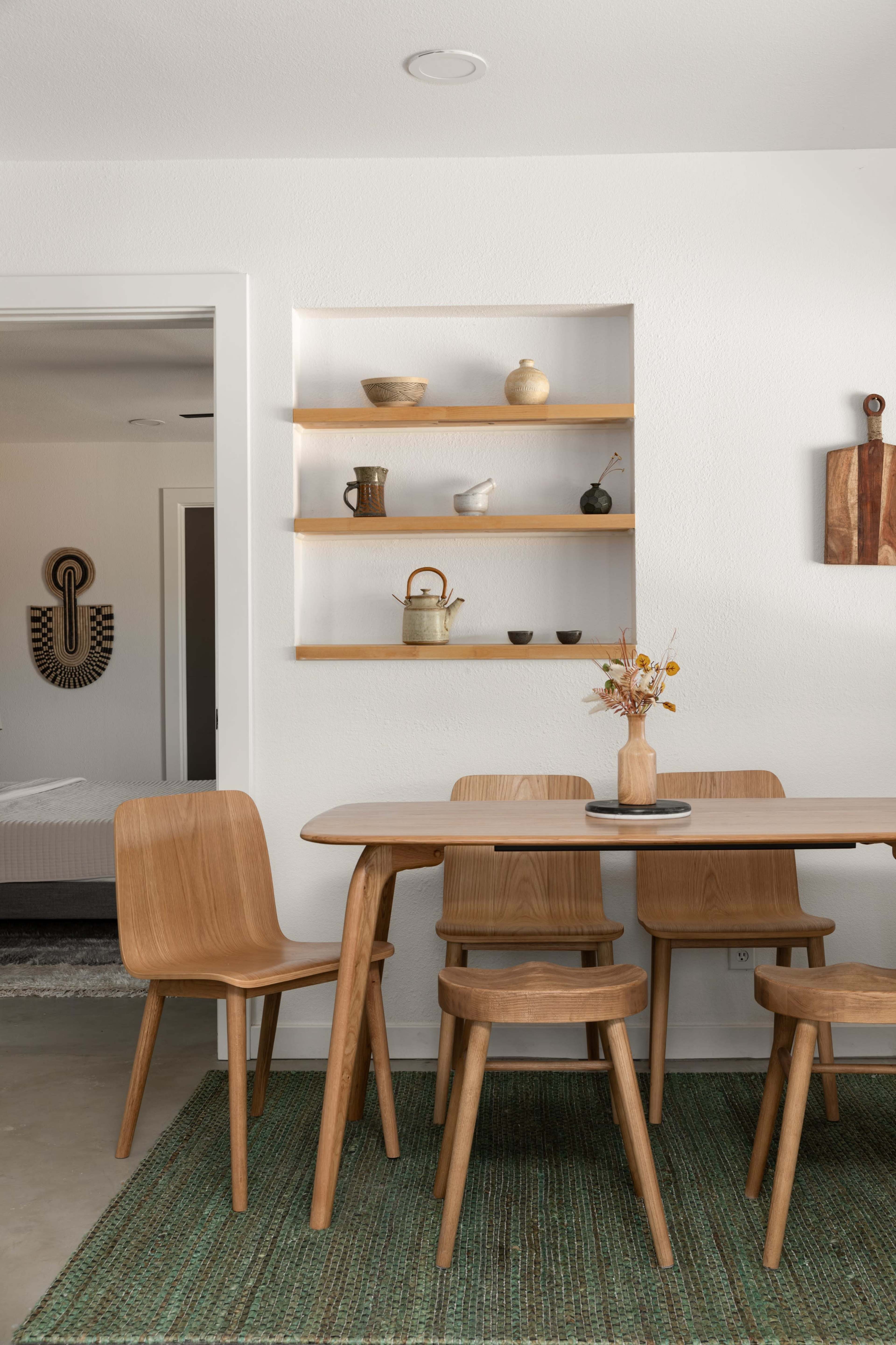 A wooden dining table with four chairs is positioned on a textured rug in a room featuring a shelf with decorative items and an adjacent doorway.