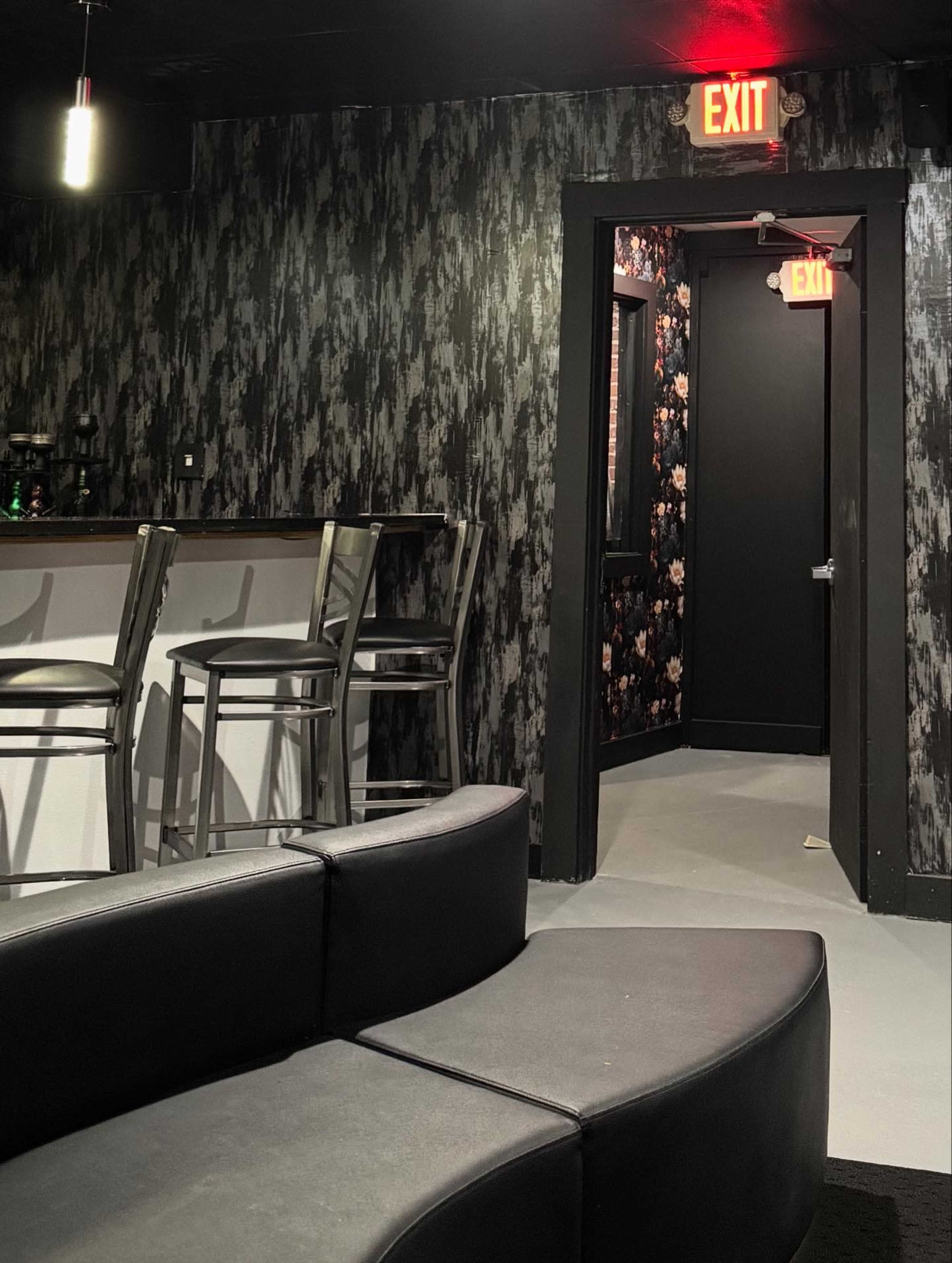 The image shows a dimly lit interior with a curved black couch in the foreground, a bar with metal stools on one side, and an exit door visible in the background.