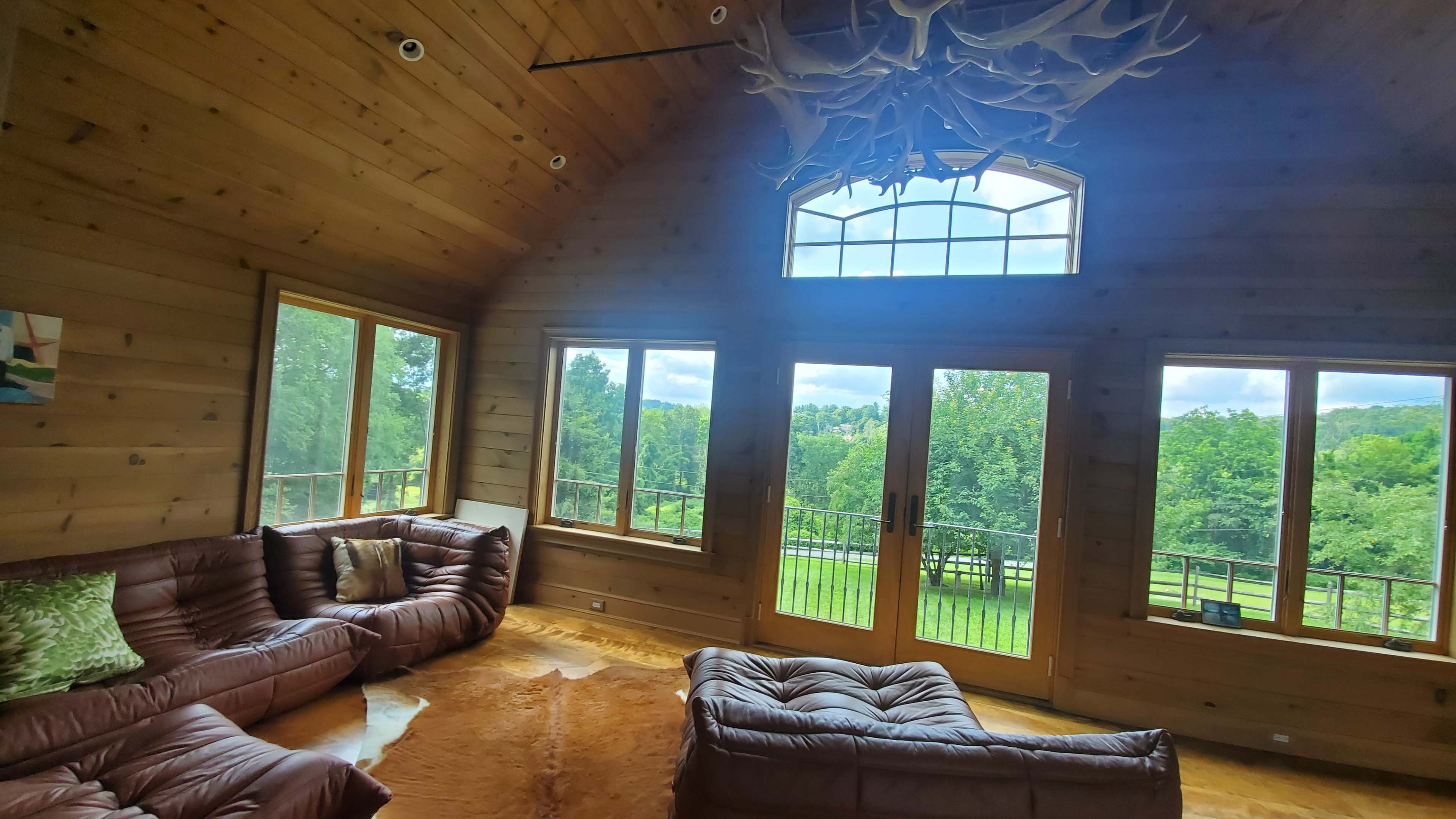 The interior of the room features wooden walls, large windows overlooking a green landscape, and leather sofas arranged around a central area rug.