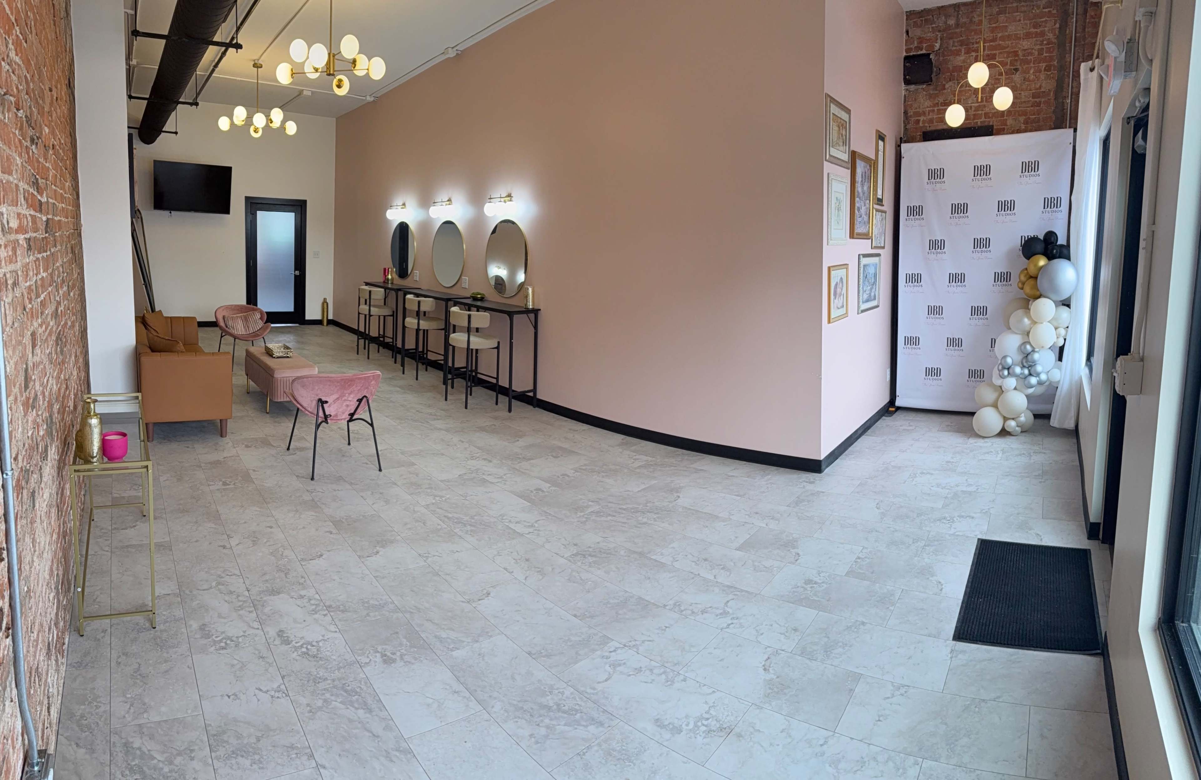 The image shows a spacious salon interior featuring pink walls, round mirrors on black vanities, a seating area with a pink couch, and a decorative wall with balloons and a logo.