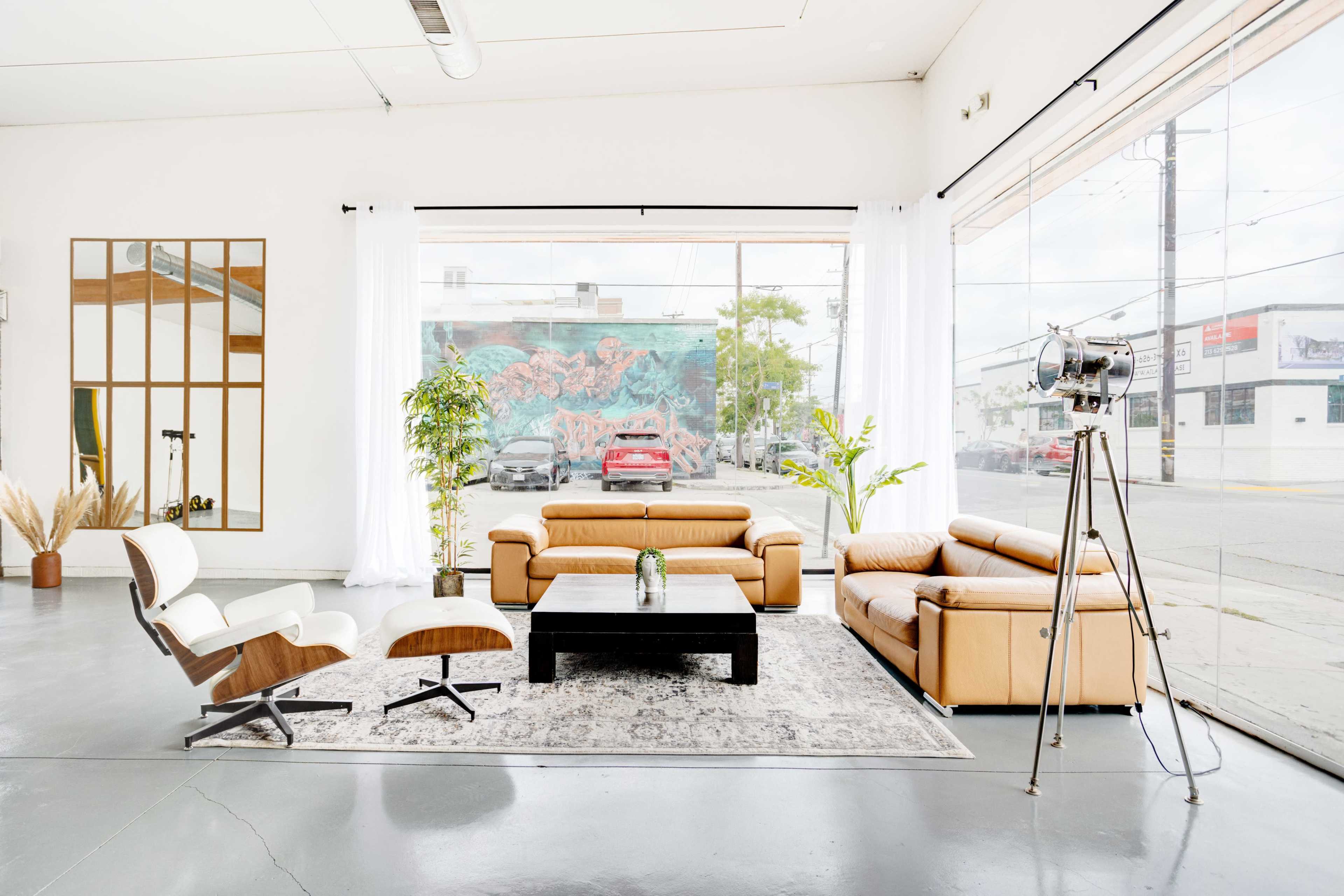 A modern living room features a tan leather sofa set, a black coffee table, a decorative plant, and a vintage floor lamp, all positioned beside large windows showcasing an outdoor mural and parked cars.