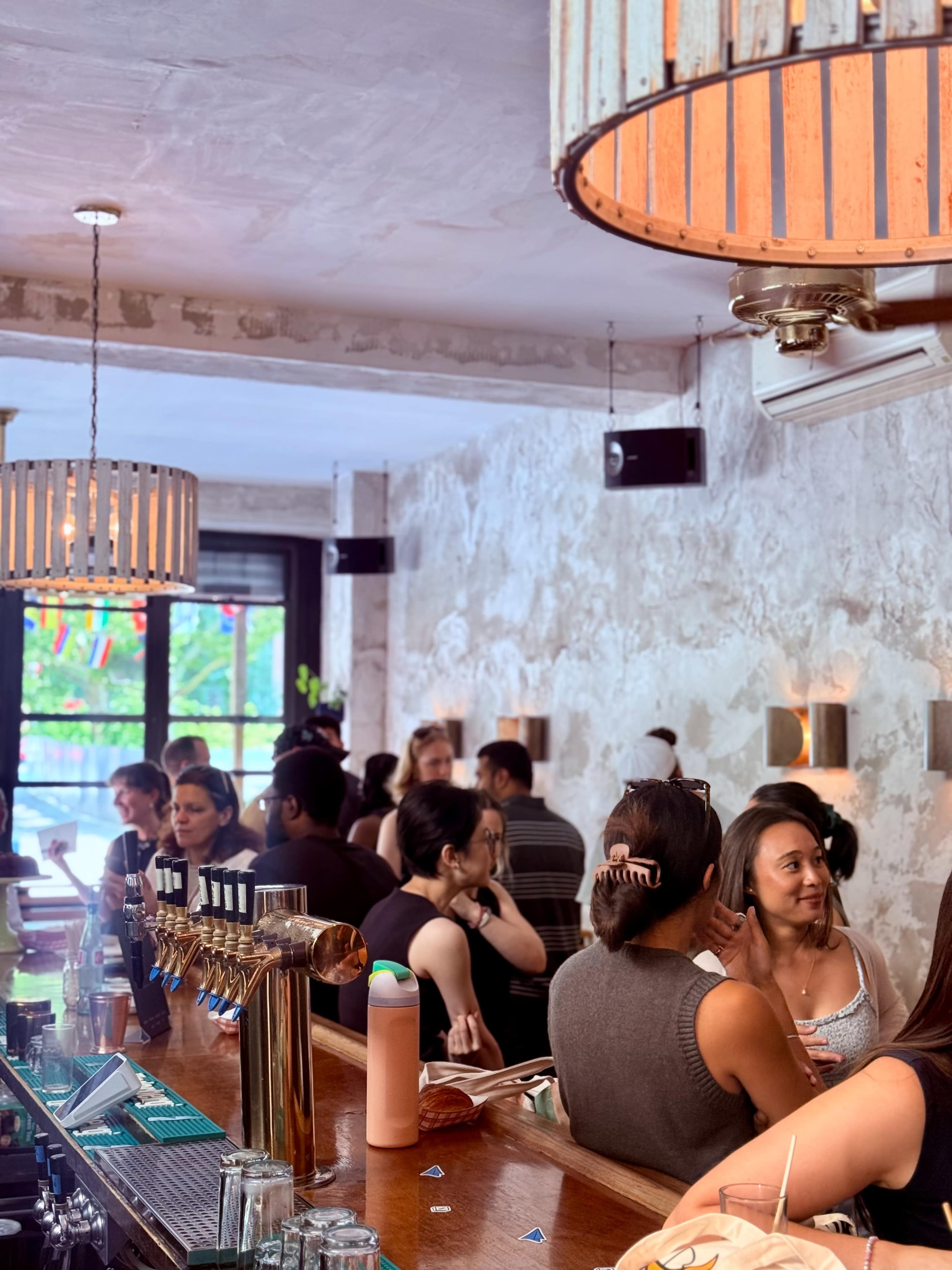 The image shows a busy bar scene with patrons gathered at the counter and socializing in a well-lit, stylish interior.