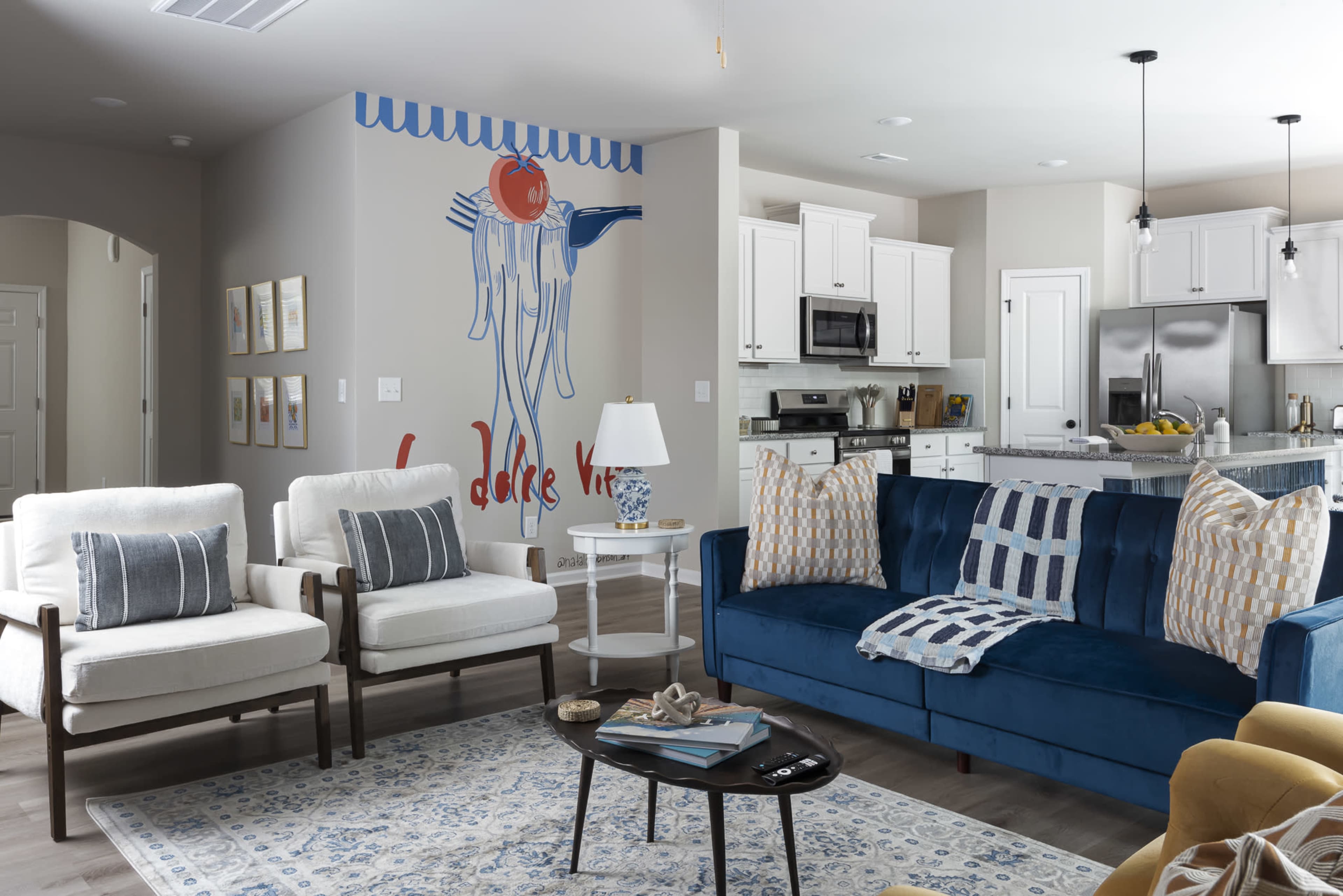 A modern living room features a blue sofa, two white armchairs, and a kitchen in the background with a large wall mural of a hanging tomato.