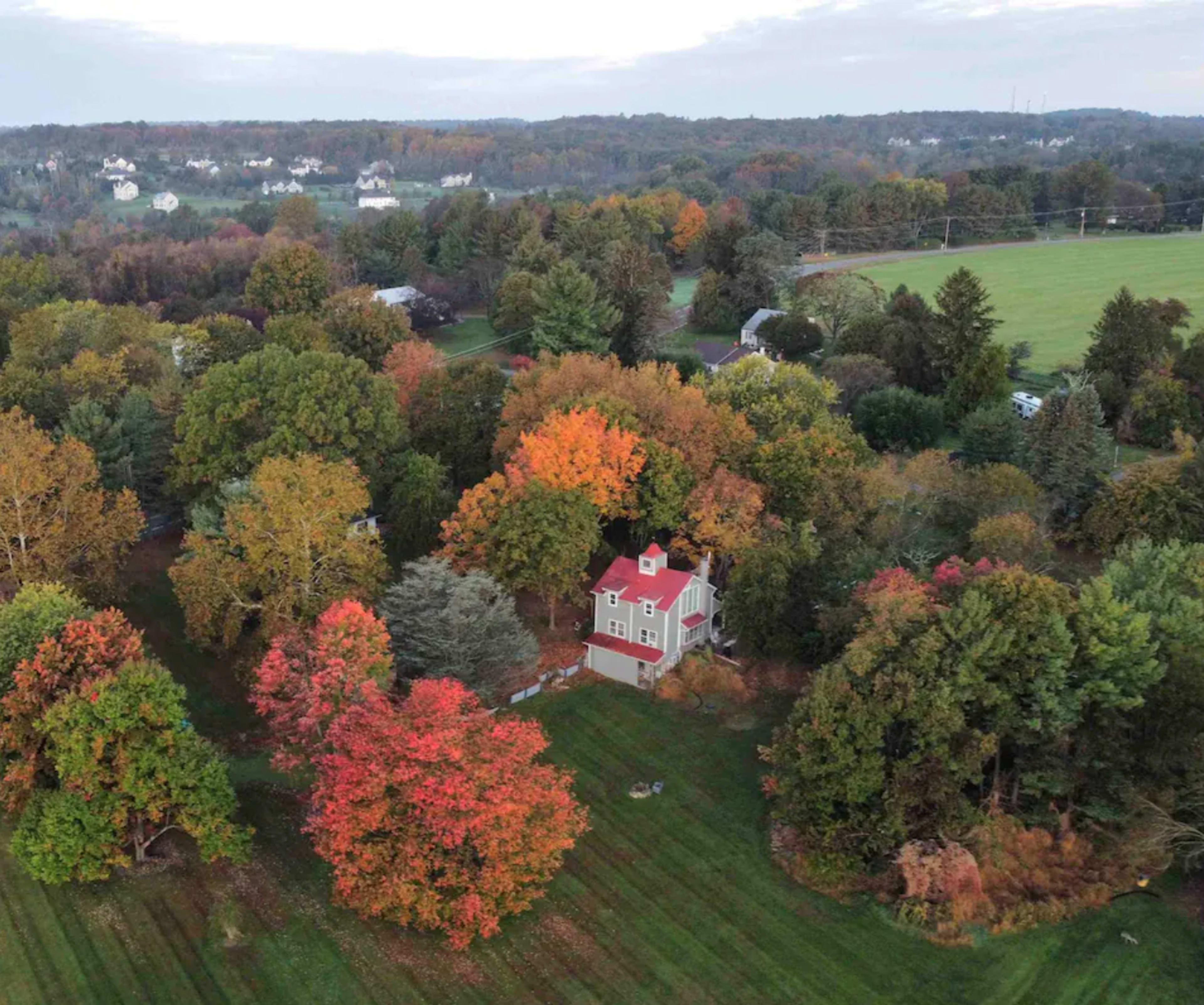 A house with a red roof is surrounded by trees displaying autumn foliage in a rural area.