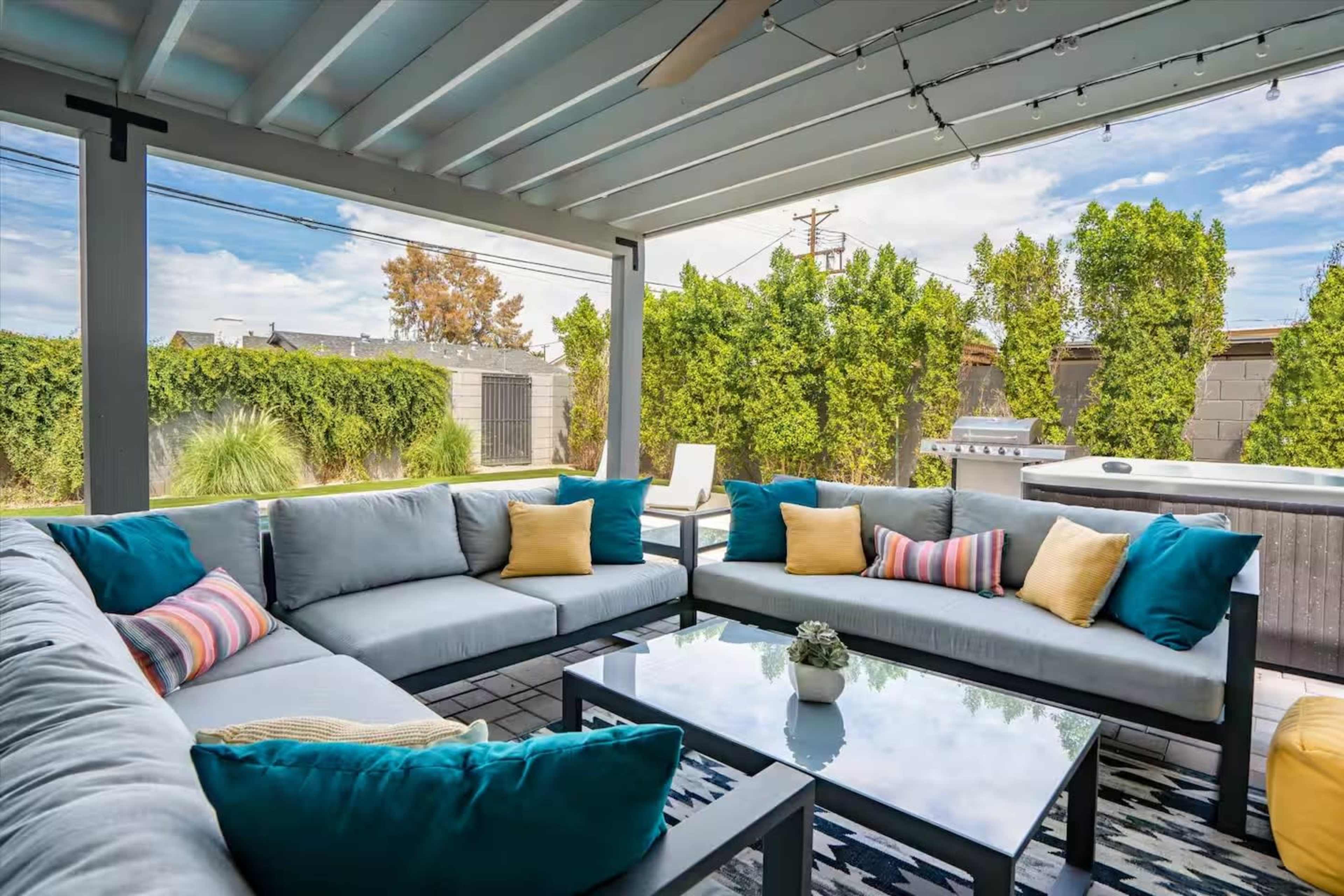 The image shows a modern outdoor seating area with gray couches adorned with colorful pillows, a glass coffee table, and a hot tub in a backyard surrounded by greenery.