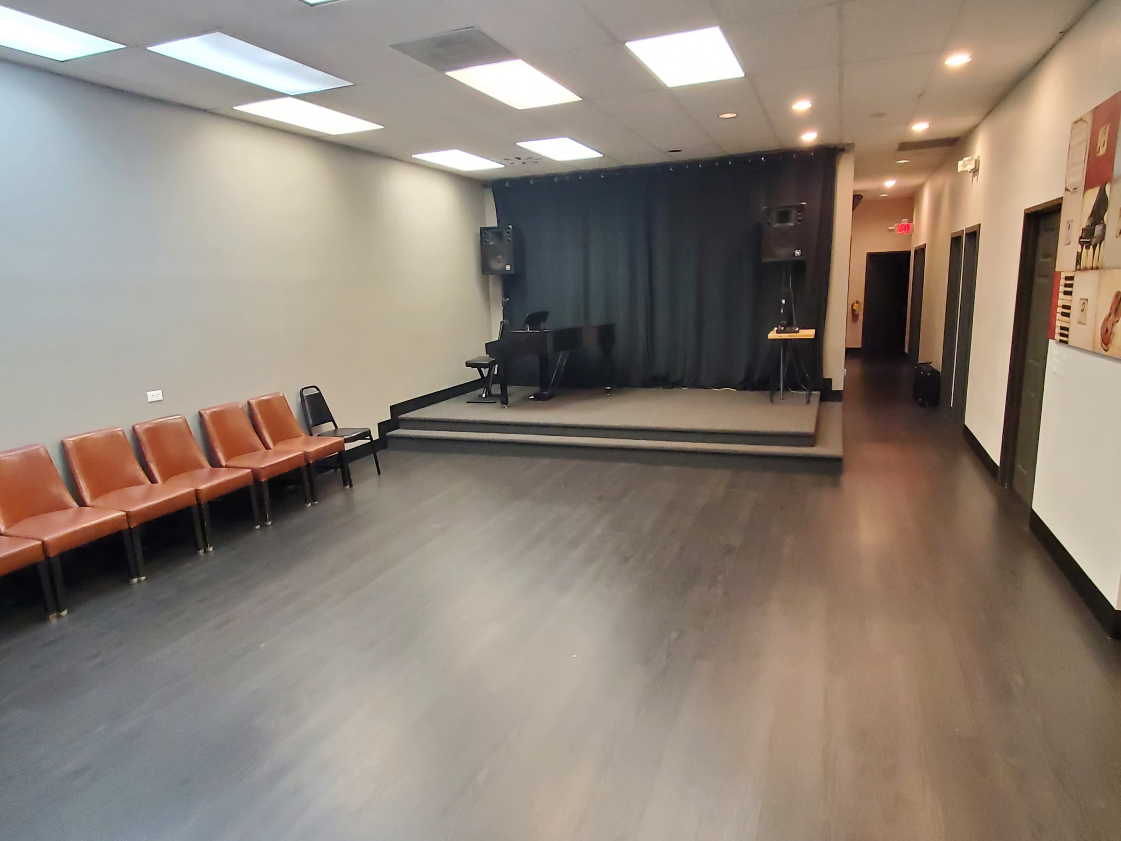 The image shows a spacious room with a small stage featuring a piano and sound equipment, along with several brown chairs lined up against one wall.
