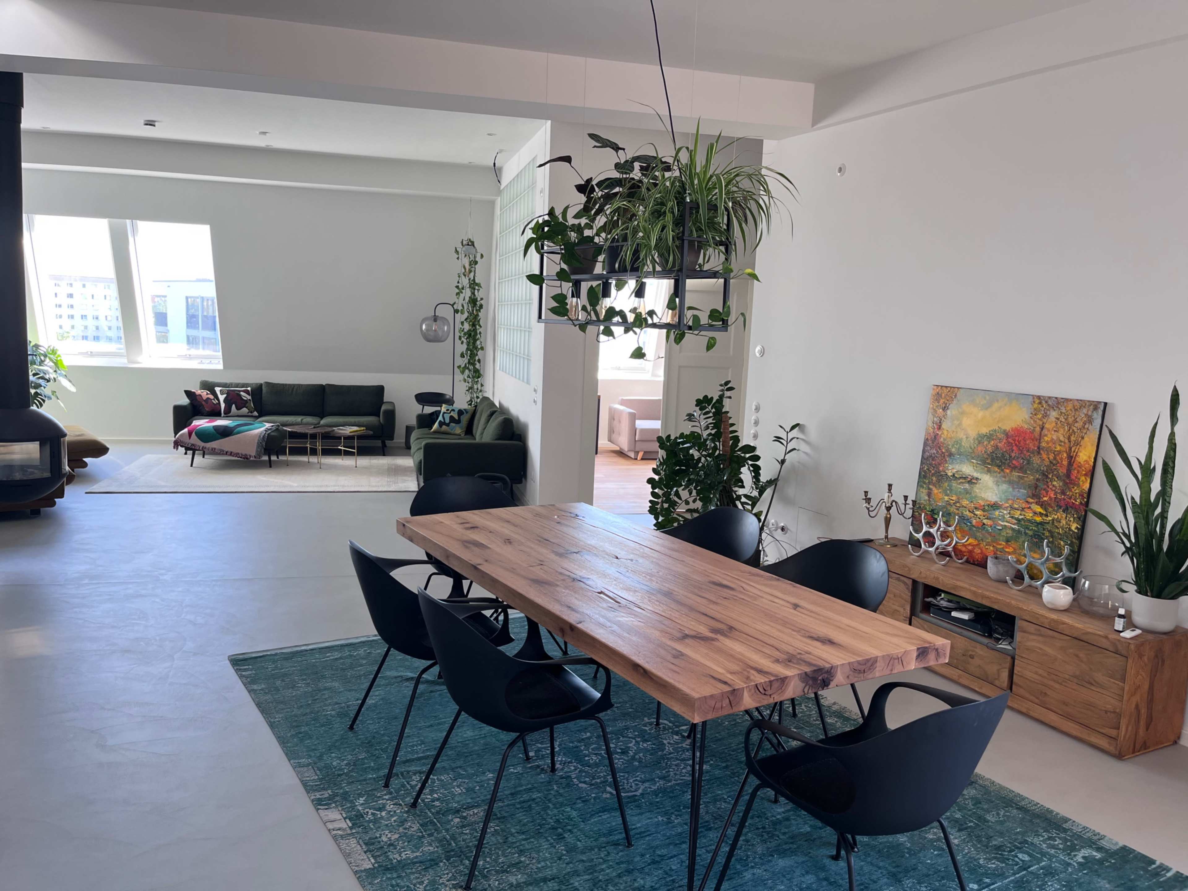 The image shows a modern, open-concept living space featuring a dining area with a large wooden table and black chairs, surrounded by plants and a colorful artwork on the wall.