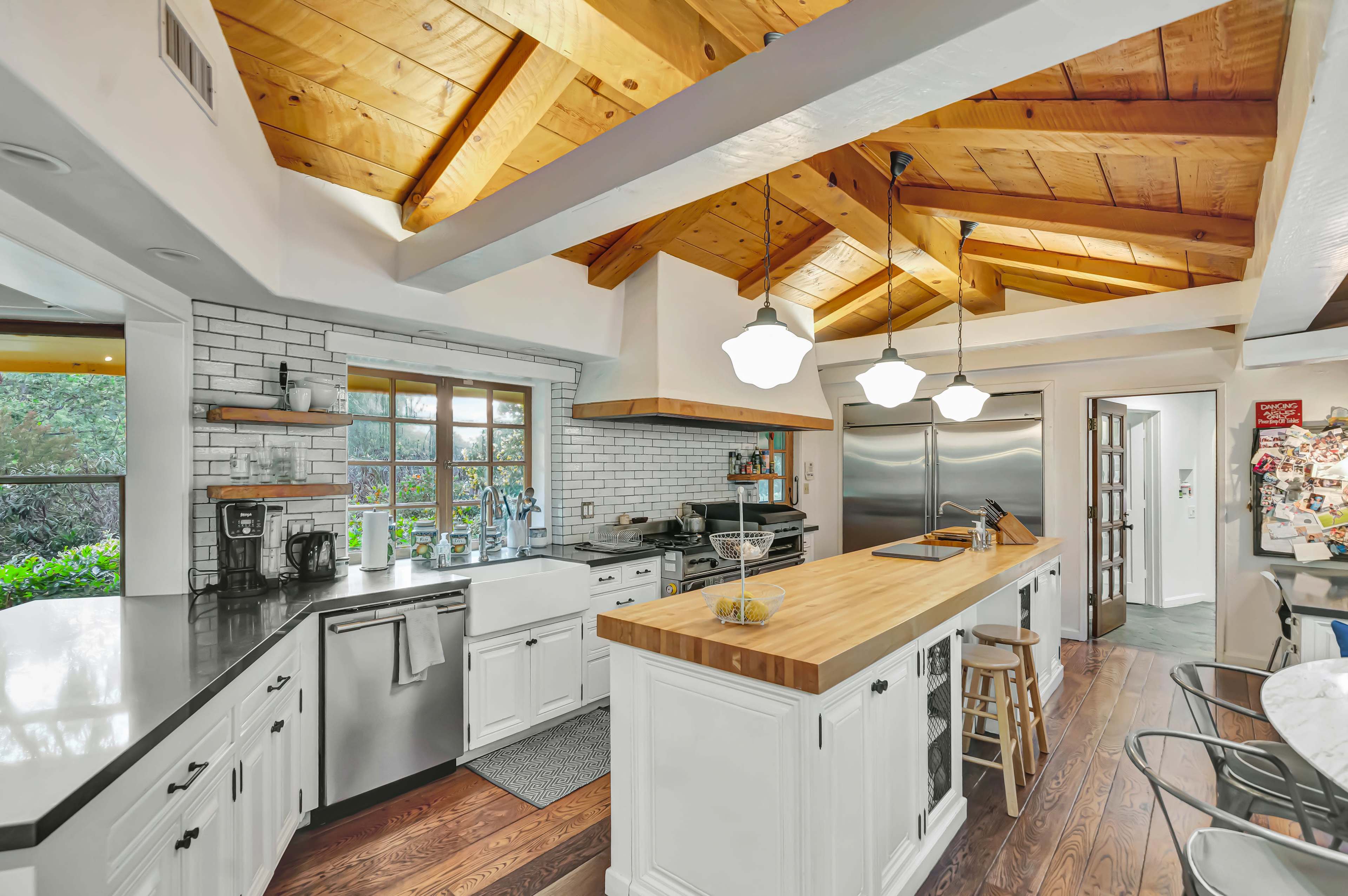 The image shows a modern kitchen featuring a large wooden island, white cabinetry, stainless steel appliances, and exposed wooden beams in the ceiling.