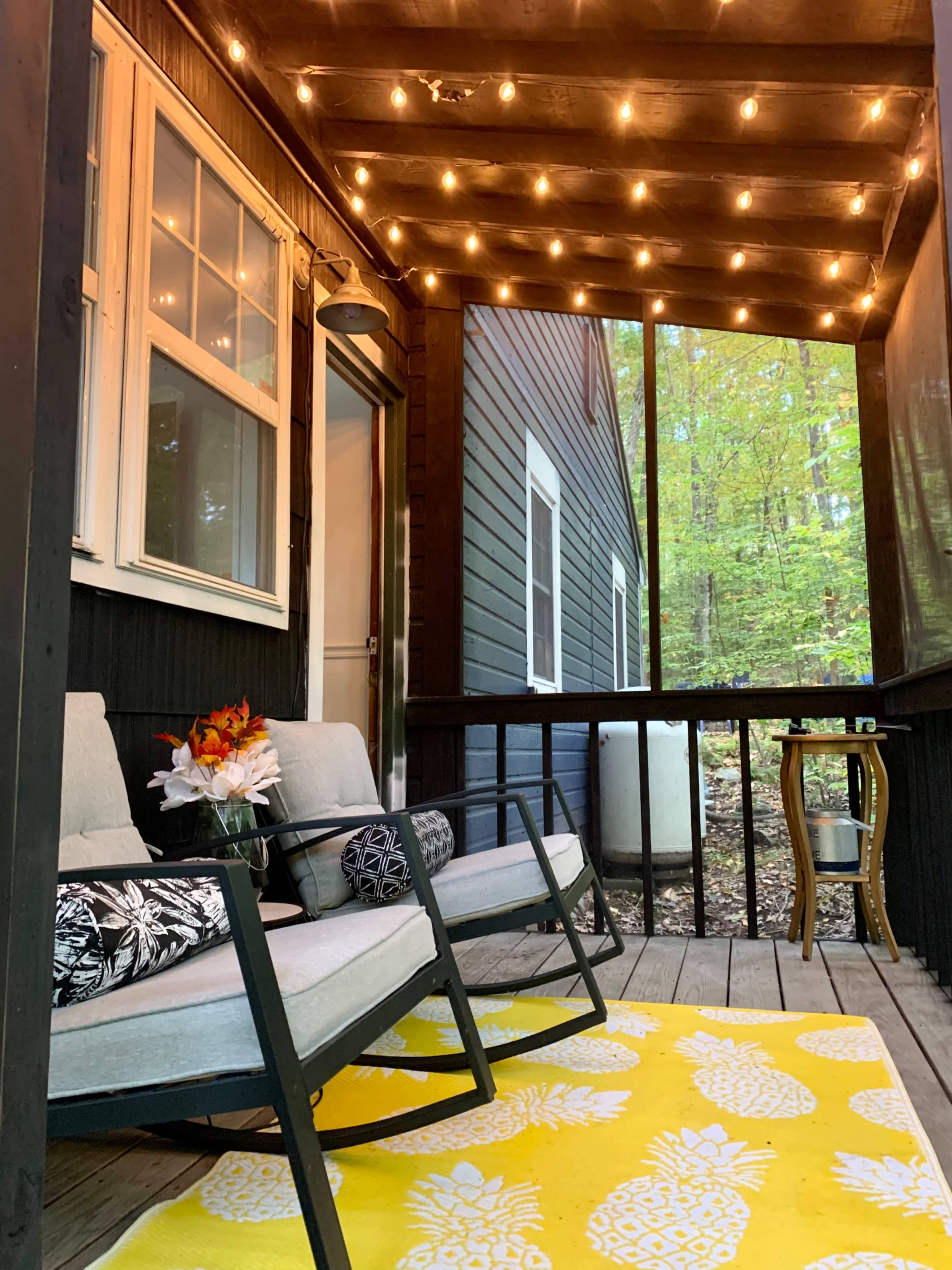 A cozy porch features two chairs, a small table, and an area rug with a pineapple design, all illuminated by string lights overhead.