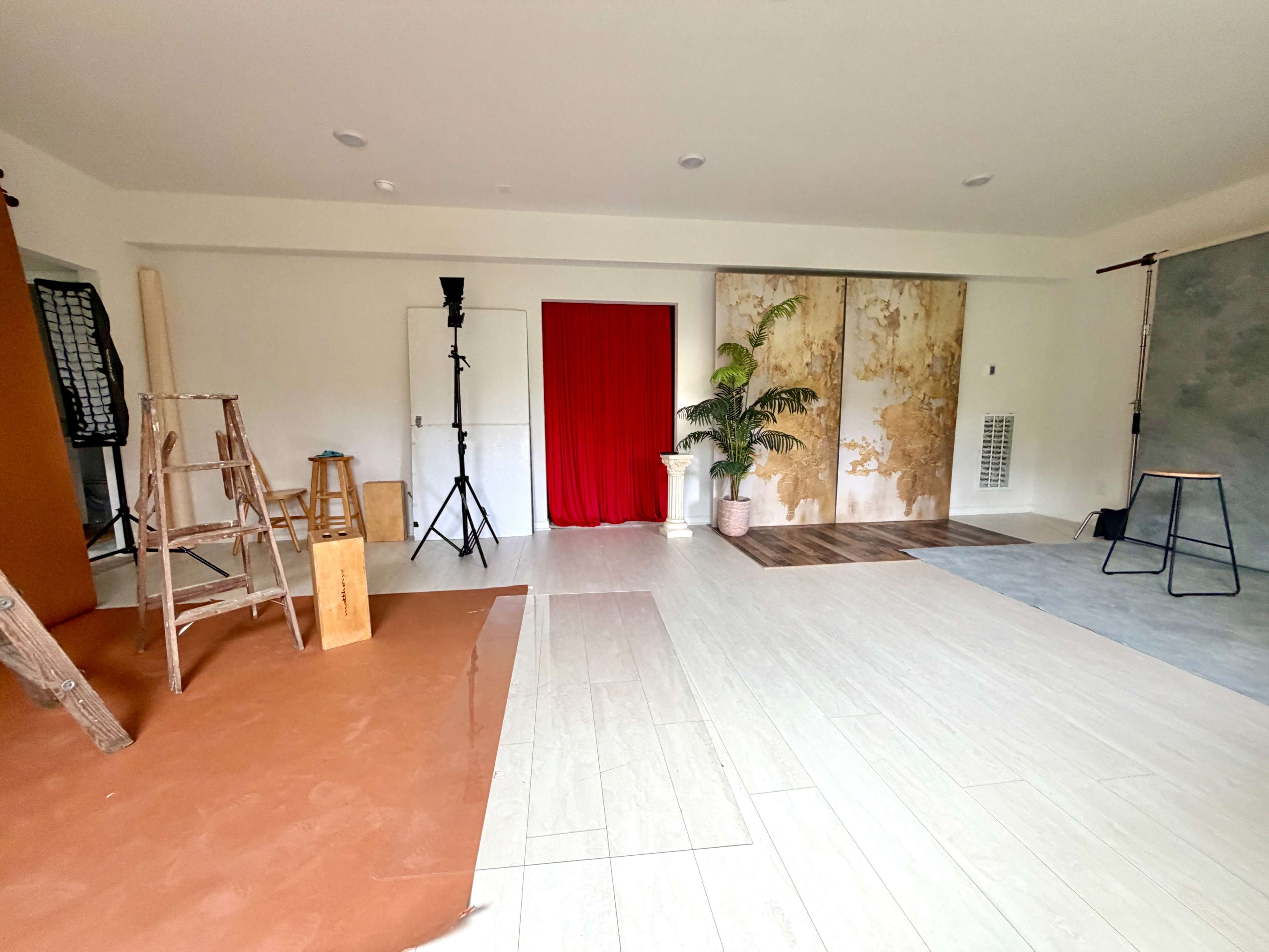 The image shows a photography studio with various backdrops, a red curtain, potted plants, and equipment set up in an open space.