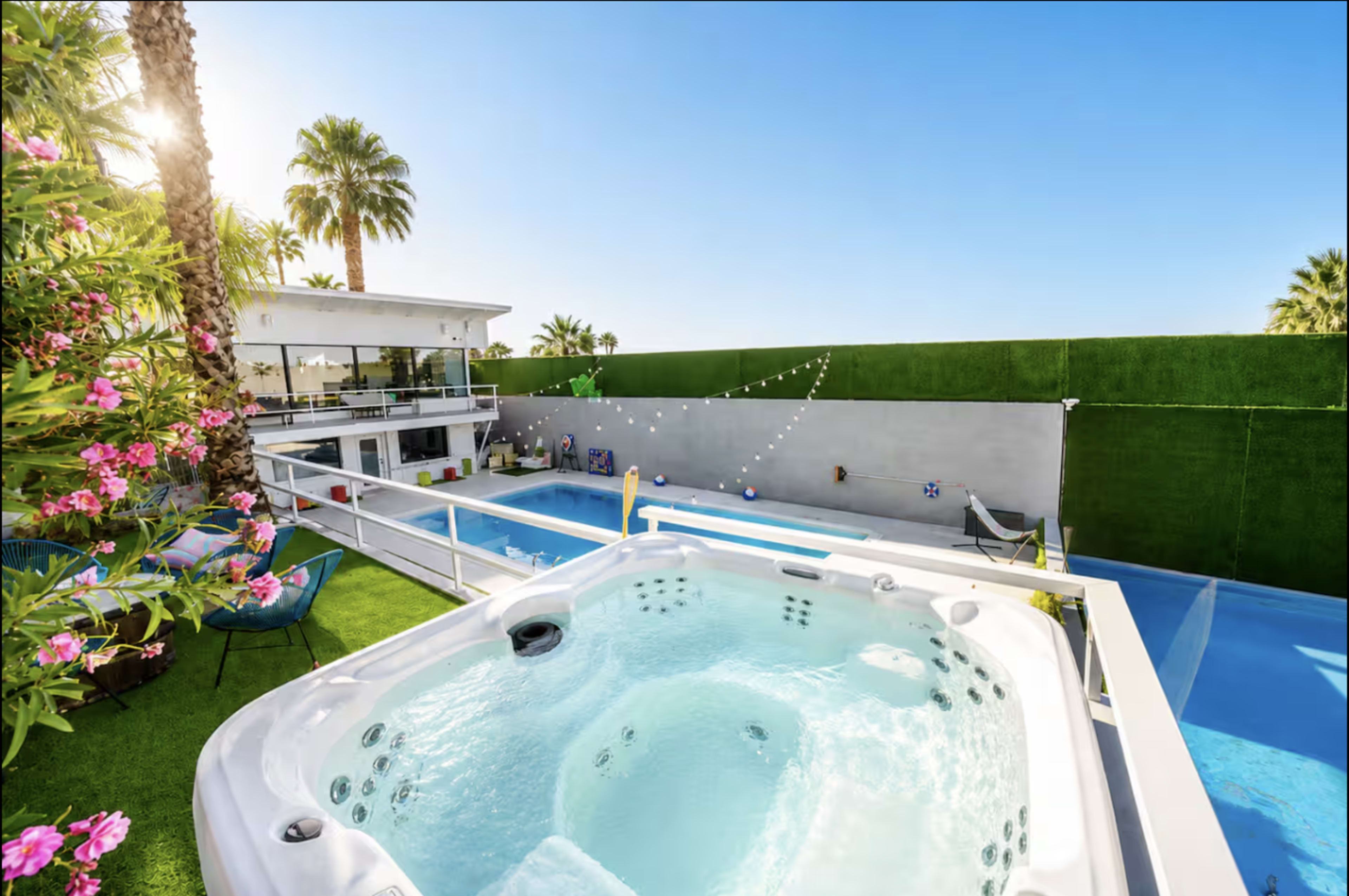 A hot tub overlooks a swimming pool surrounded by palm trees and a modern house in a sunny outdoor setting.