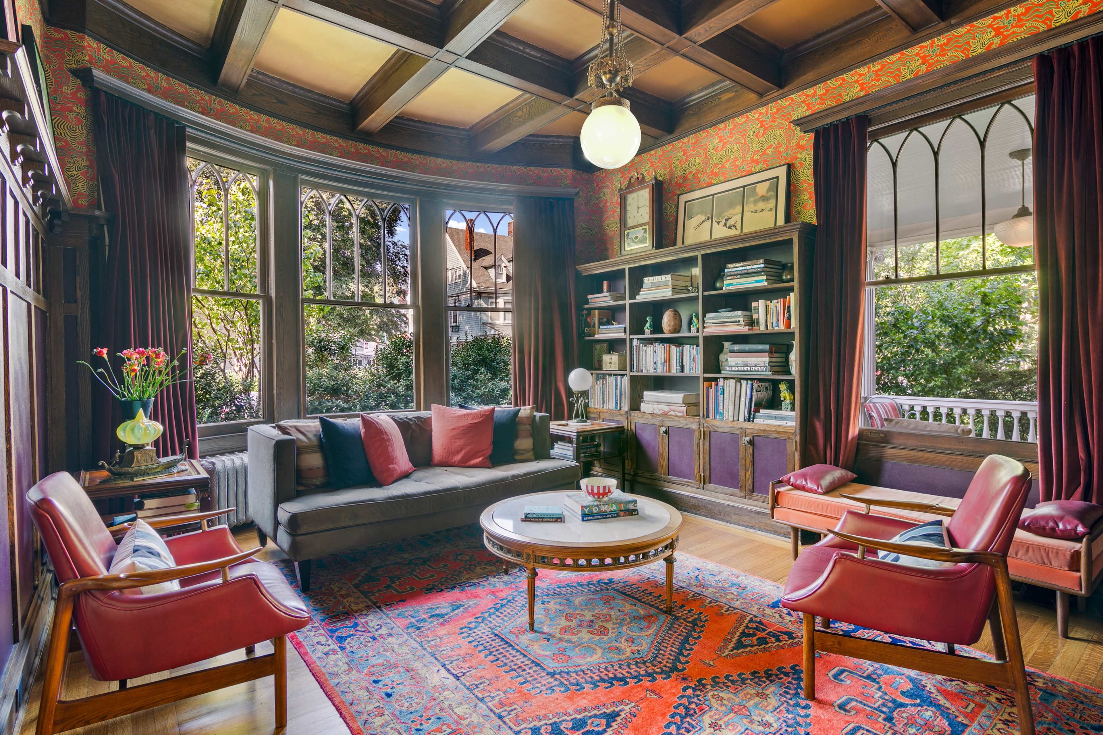 The image shows a cozy living room with a large window, a gray sofa adorned with red pillows, a round coffee table, and dark wooden shelves filled with books.