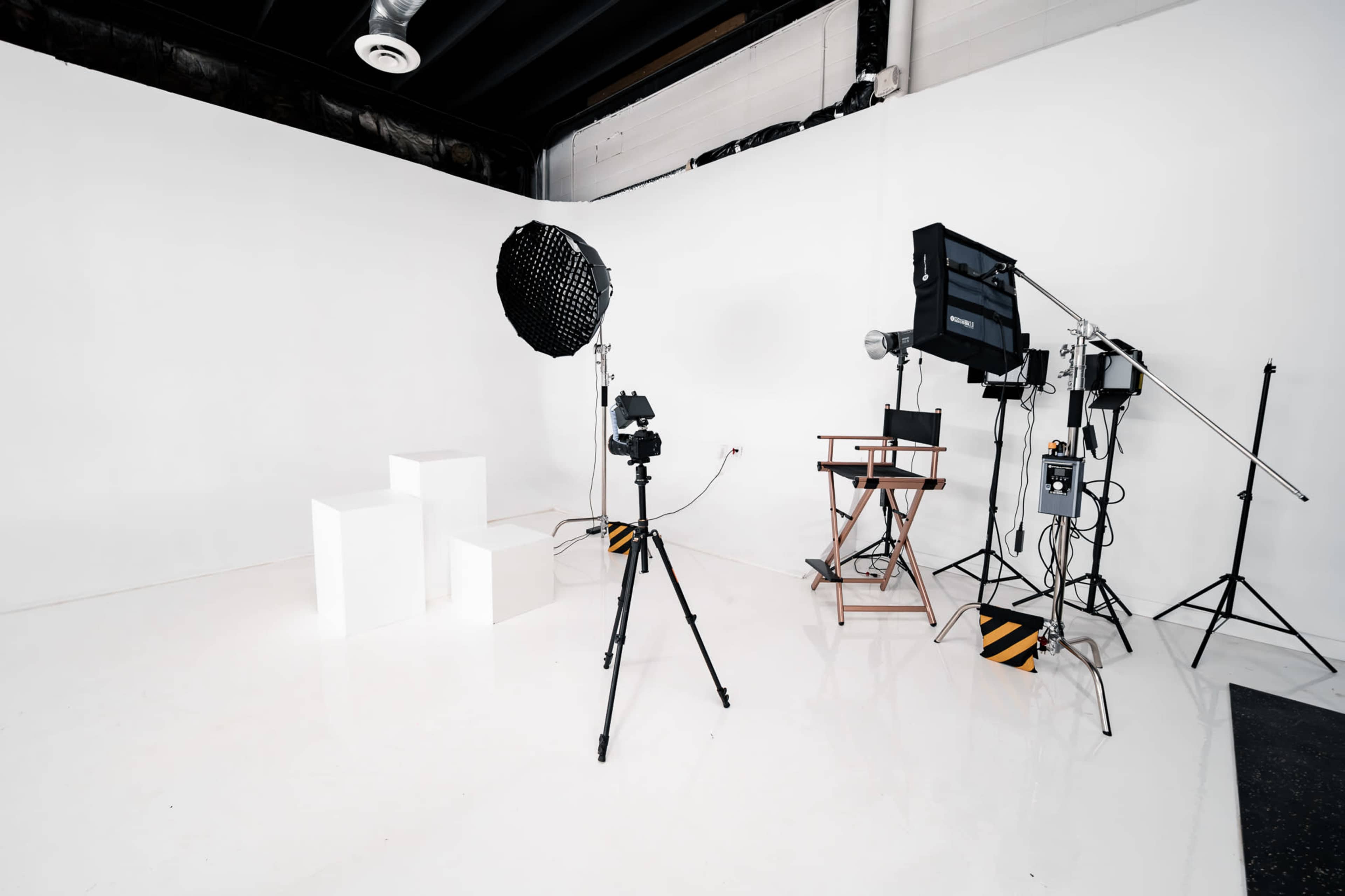 The image shows a photography studio with white walls, a shiny floor, and various lighting equipment set up around three white blocks and a director's chair.