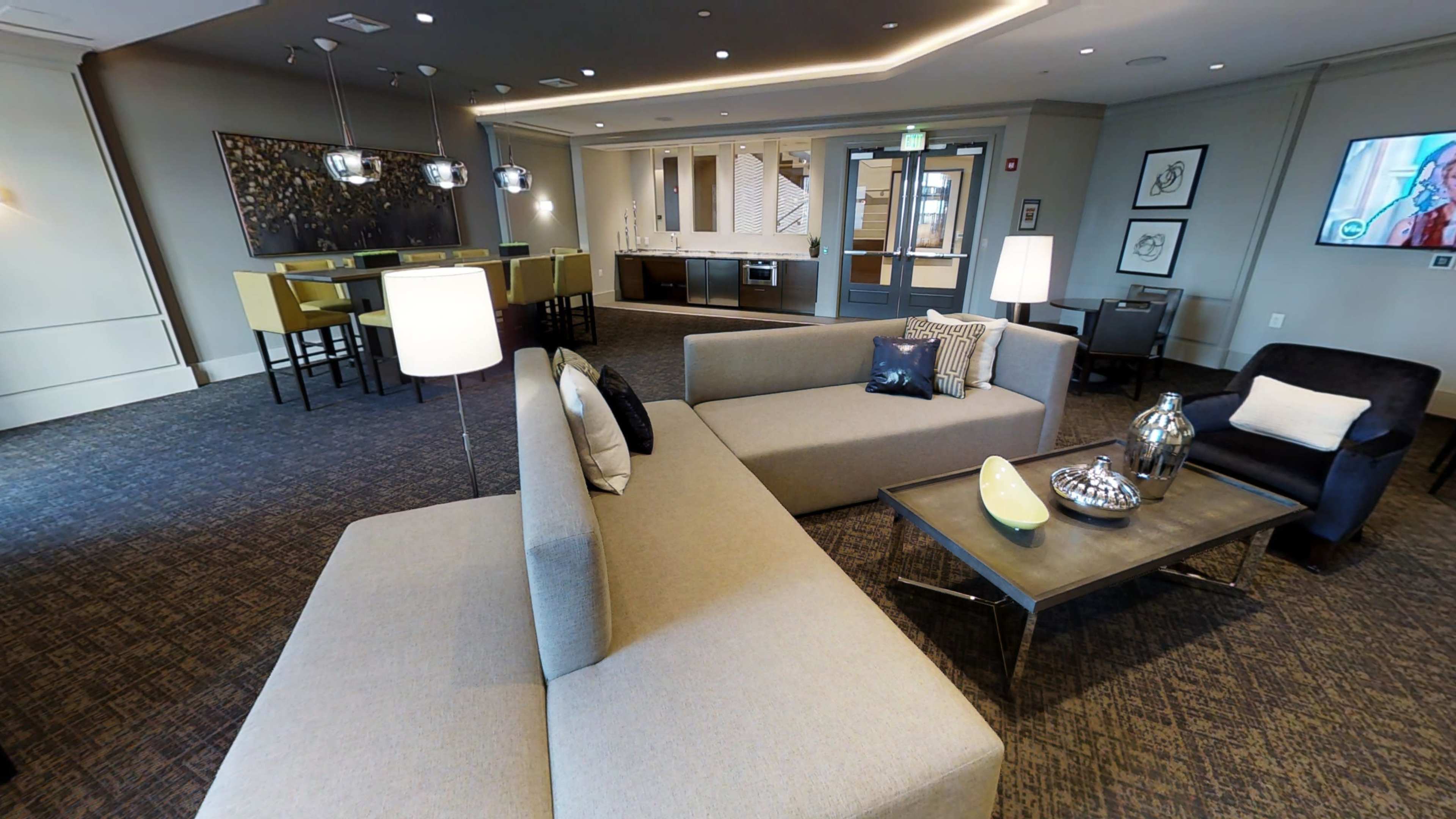 The image shows a modern lounge area featuring gray sofas, a coffee table, and a dining space in the background with a kitchen setup.