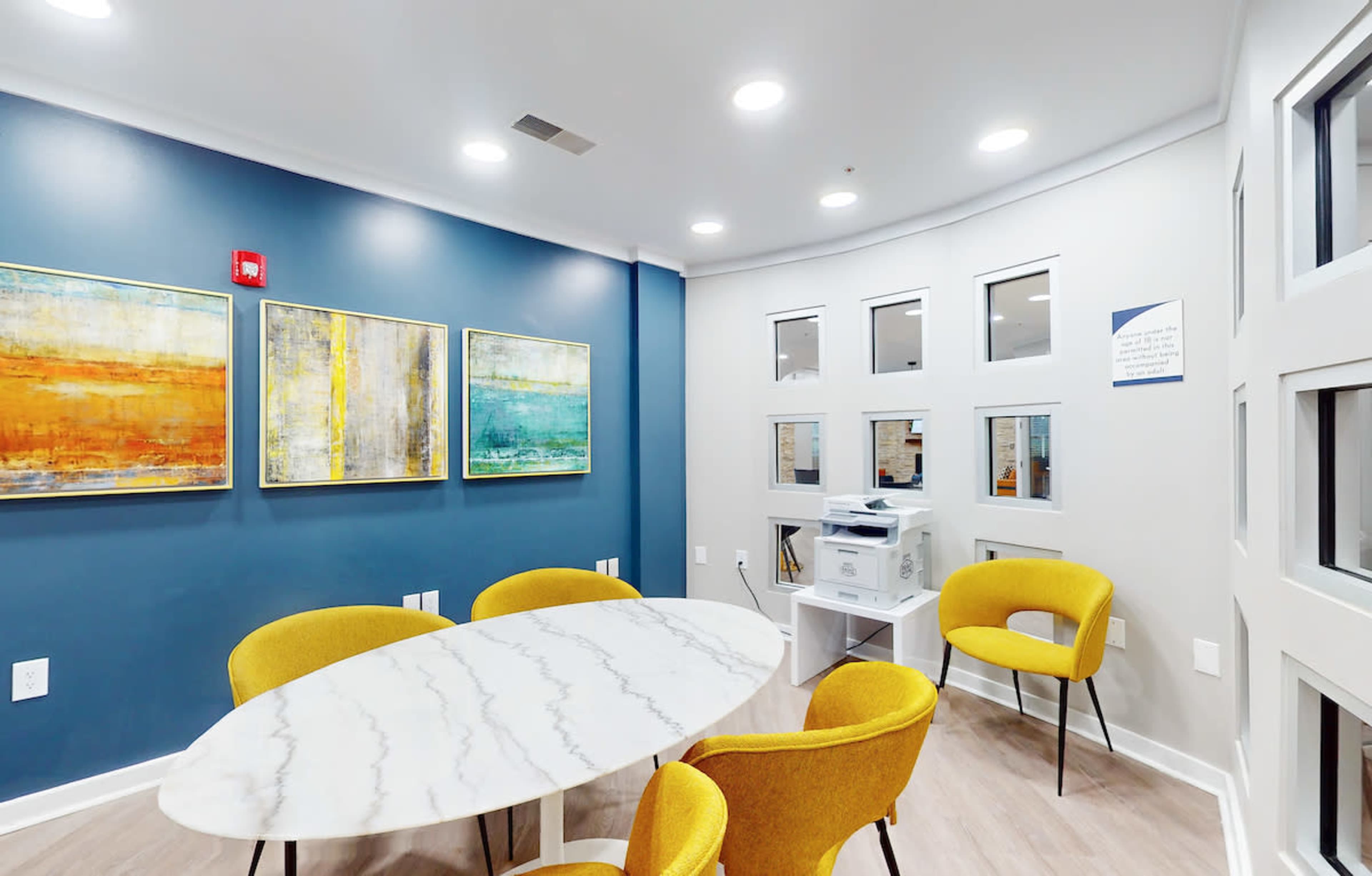 A small meeting room features a marble-top table surrounded by yellow chairs, with blue walls adorned with three abstract paintings and a printer in the corner.
