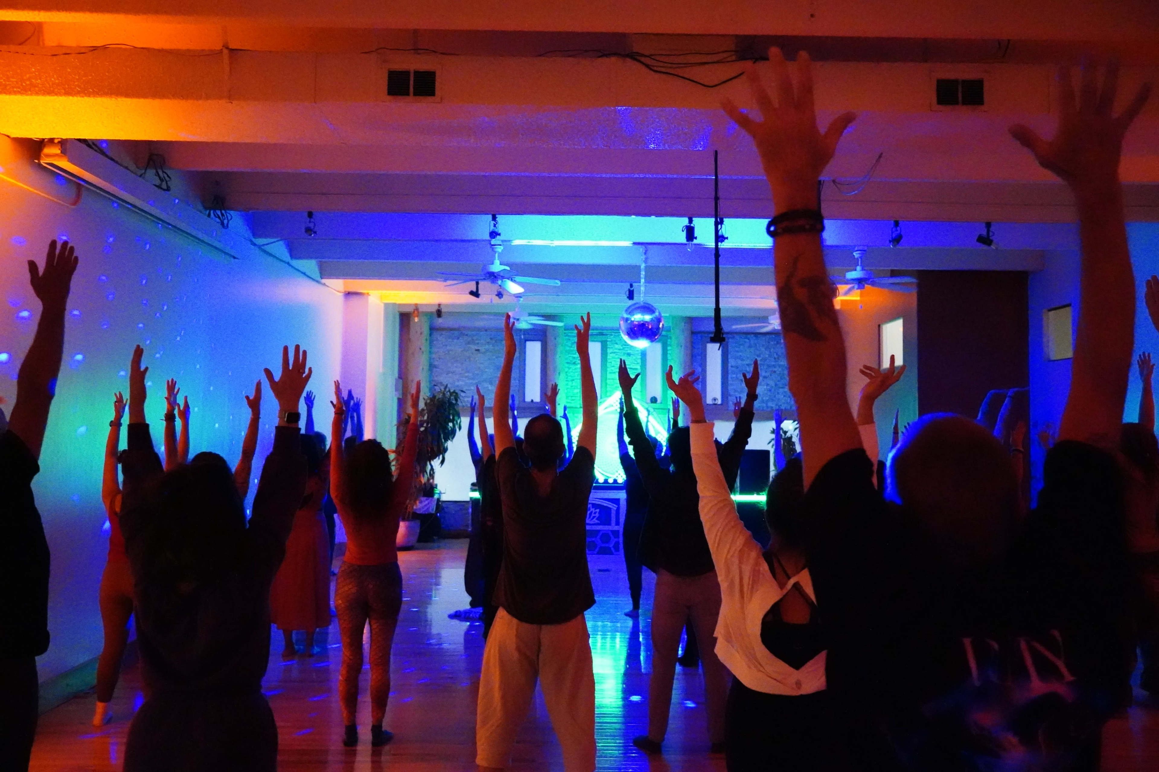 A group of people stands with their arms raised in a brightly lit dance studio filled with colorful lights.