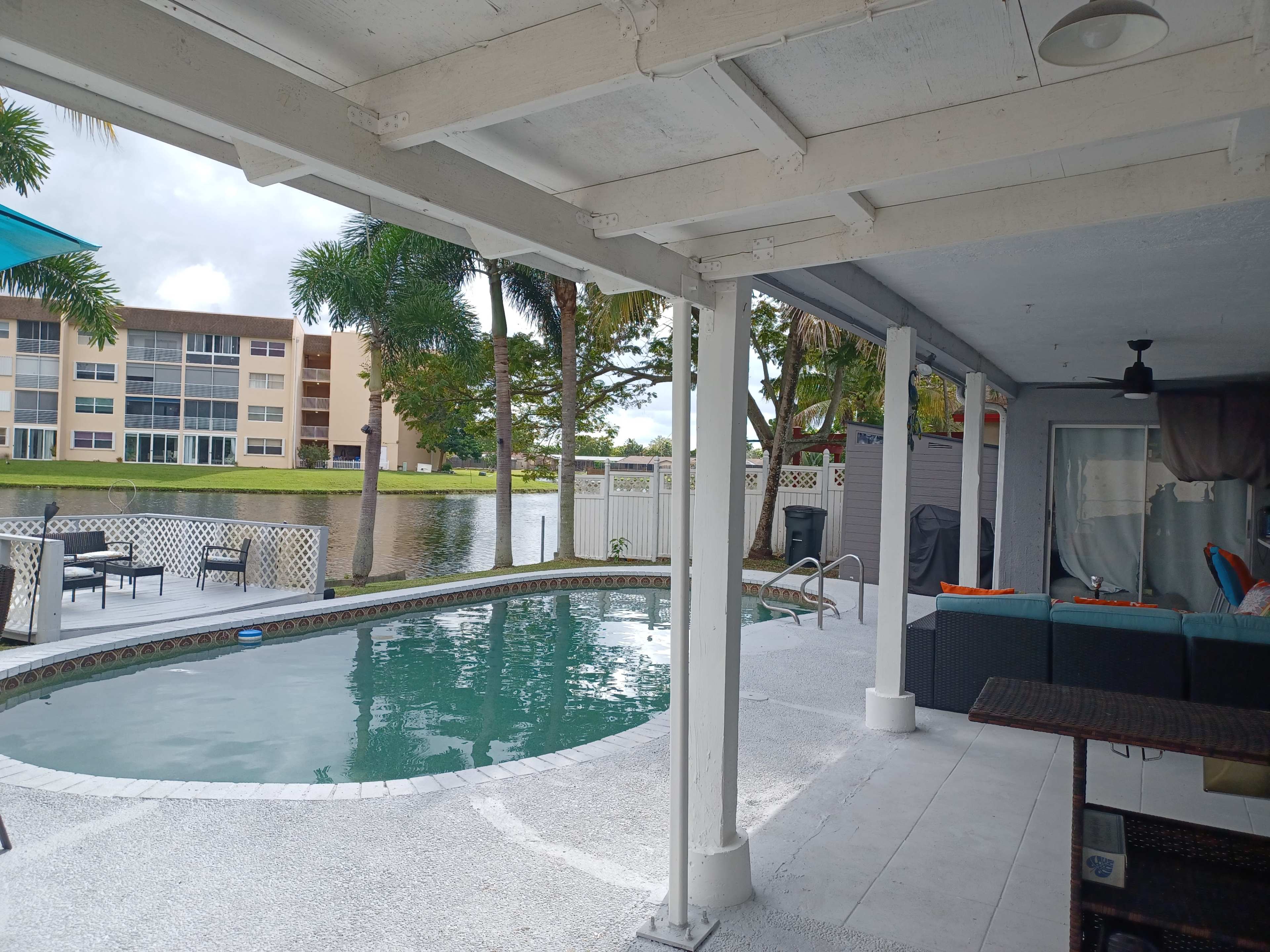 The image shows a backyard scene featuring a pool, adjacent seating area, and a view of a waterfront with nearby buildings.