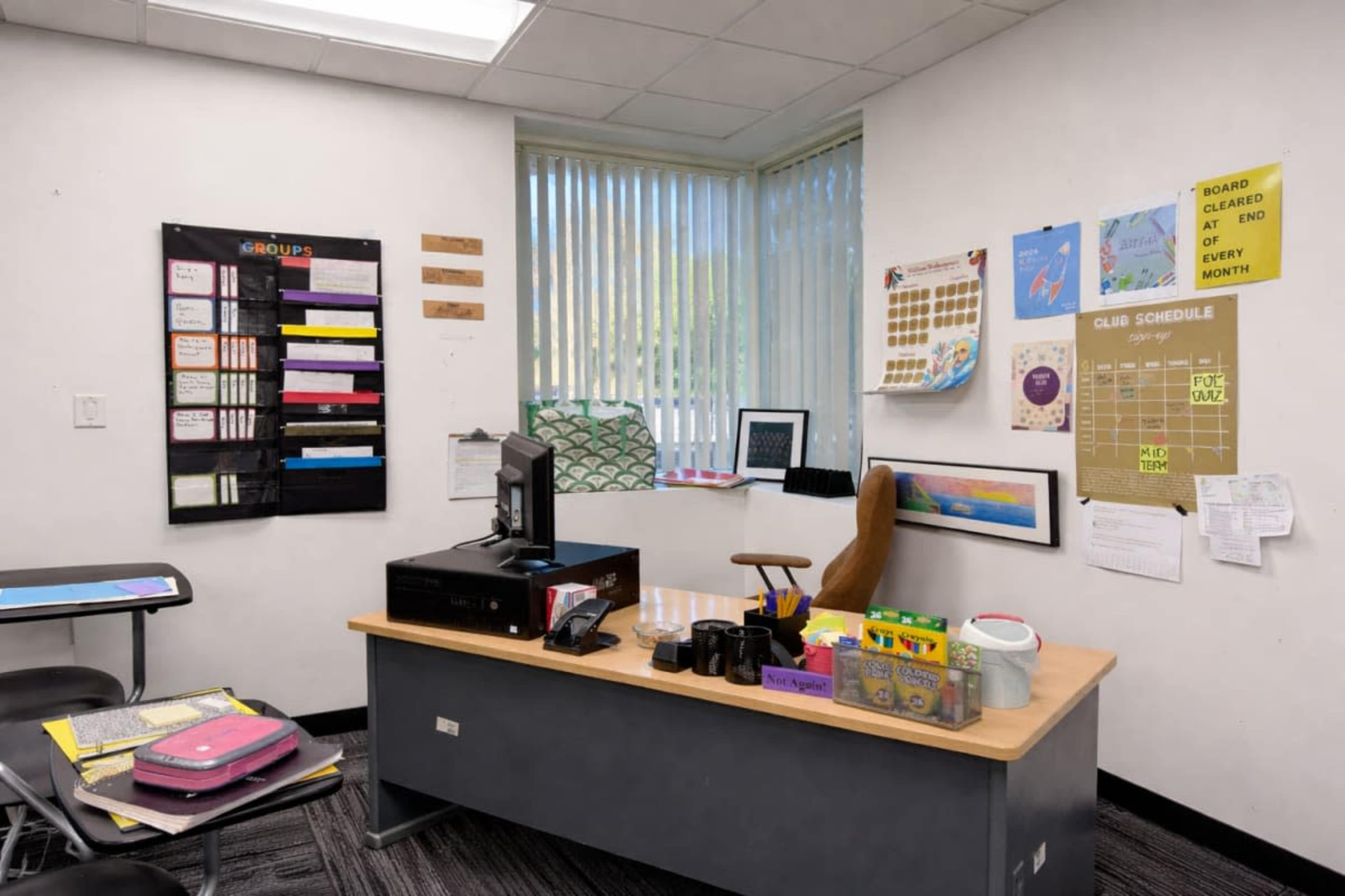 The image depicts a classroom with a teacher's desk, a computer, and various educational materials pinned on the walls.