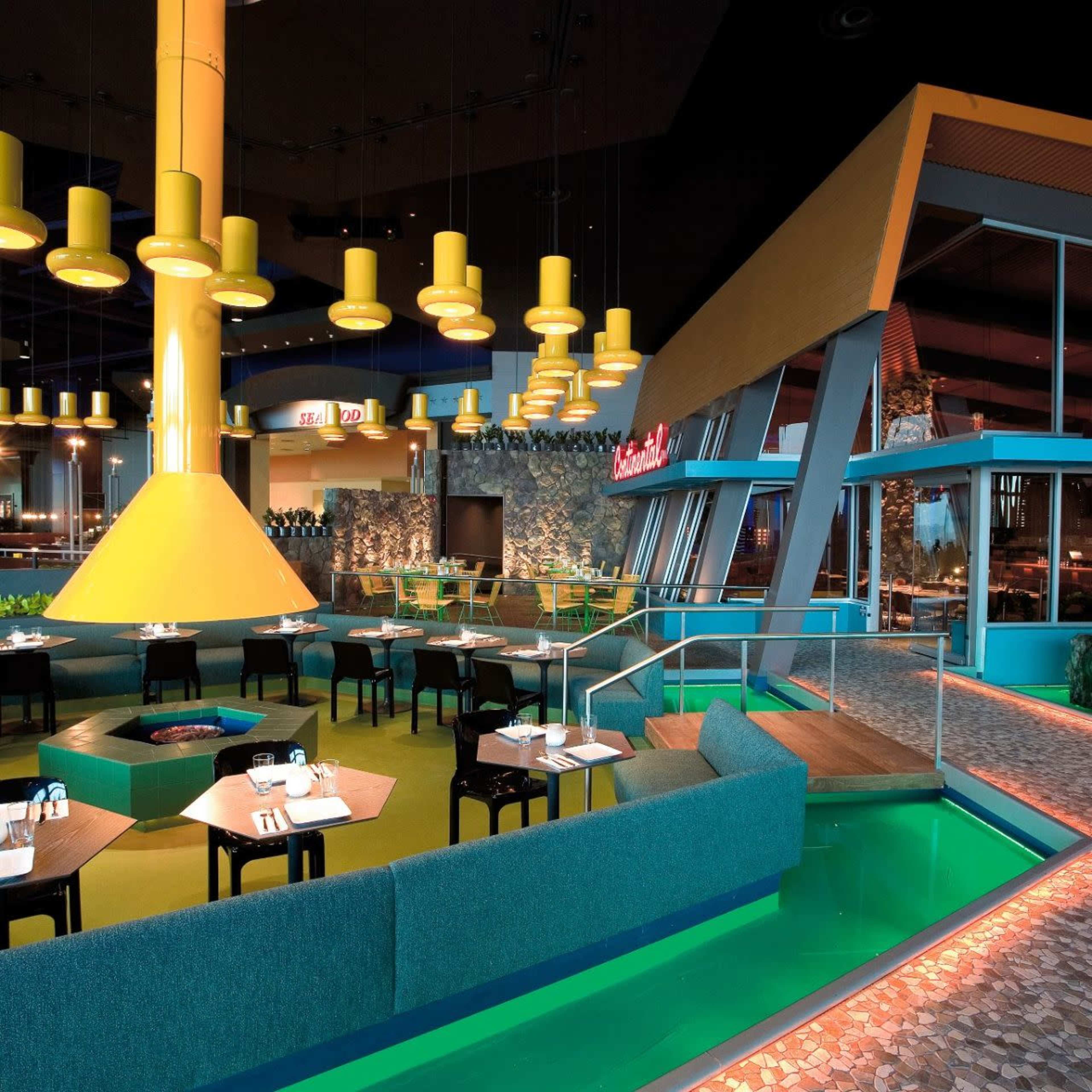 A modern restaurant interior featuring a colorful design with yellow hanging lights, a green floor, and a large central column.