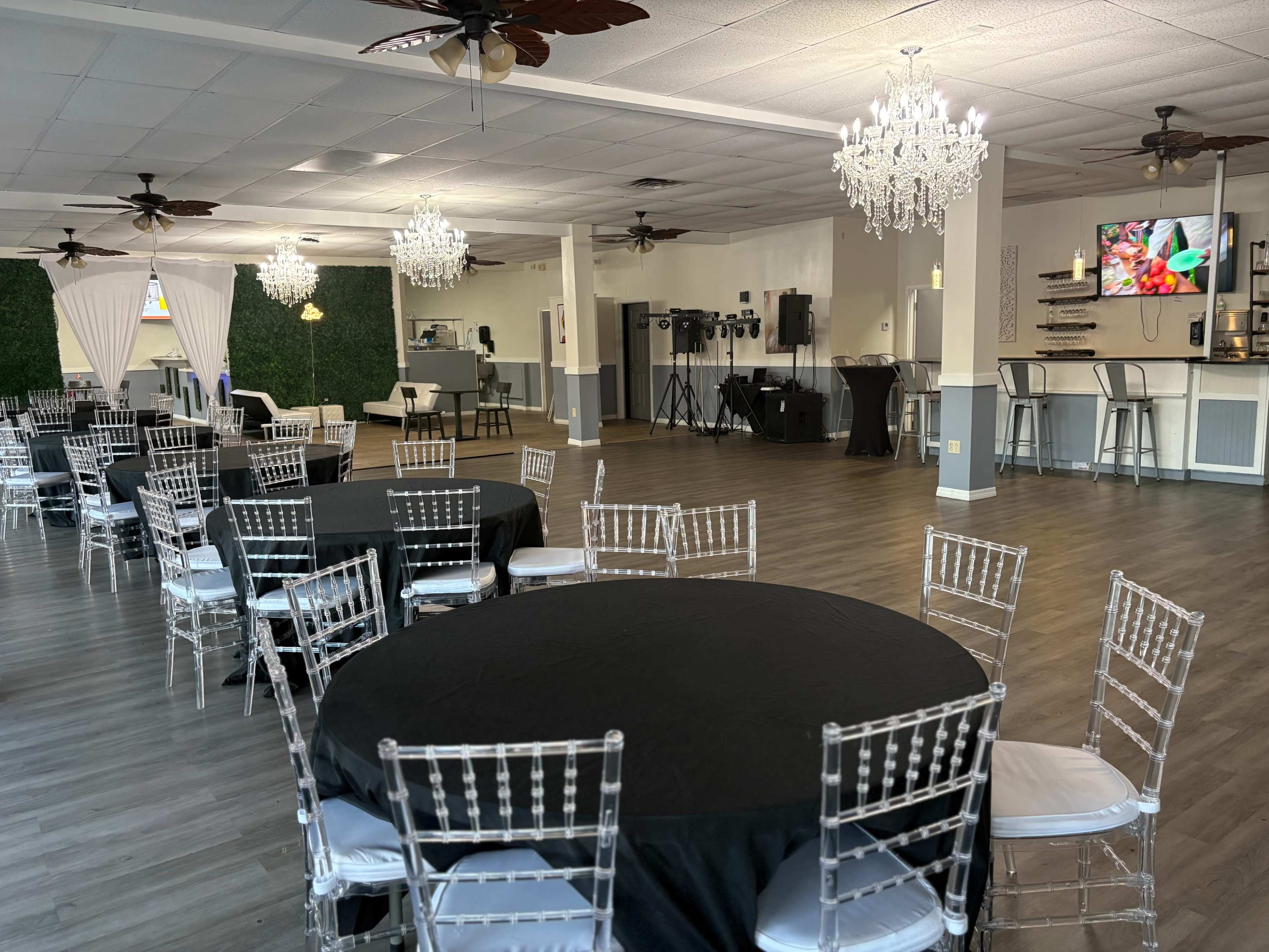 The image shows a spacious event hall with several round tables covered in black tablecloths, surrounded by transparent chairs, and featuring chandeliers and a bar area in the background.