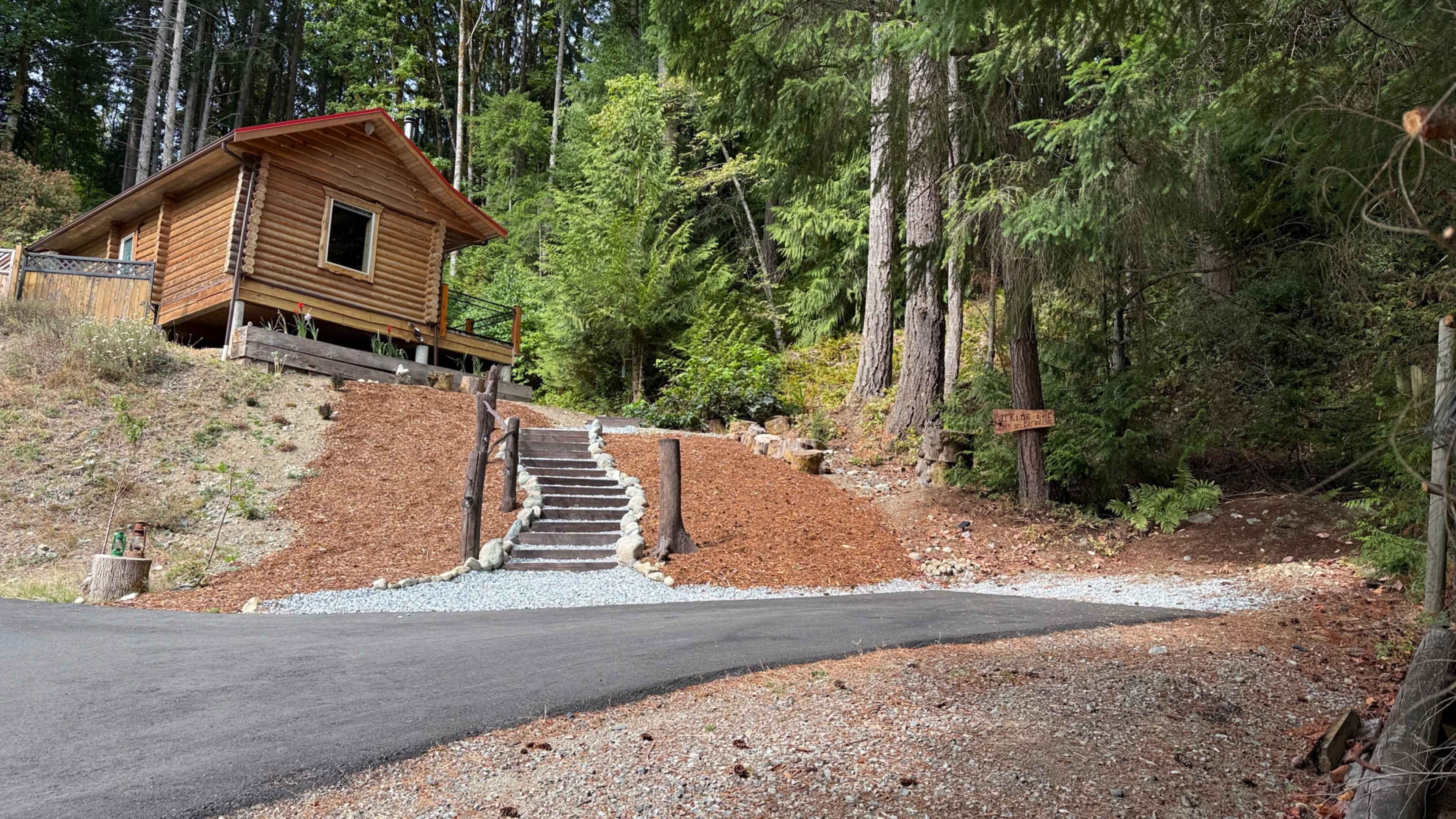 A wooden cabin sits at the edge of a forested area, with a gravel path leading up a set of steps to its entrance.