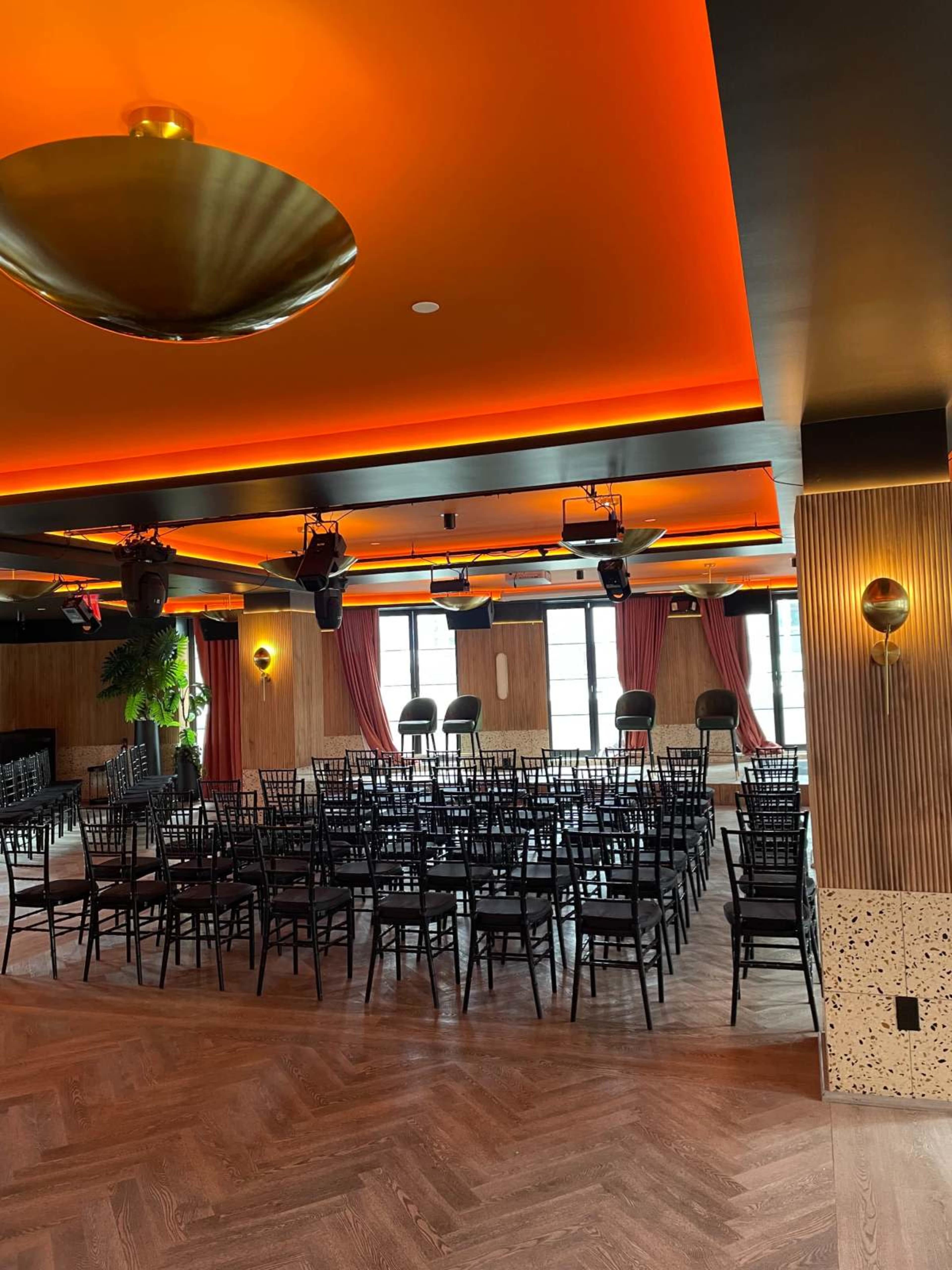 The image shows a spacious event venue set up with rows of black chairs, adorned with orange lighting and contemporary decor.