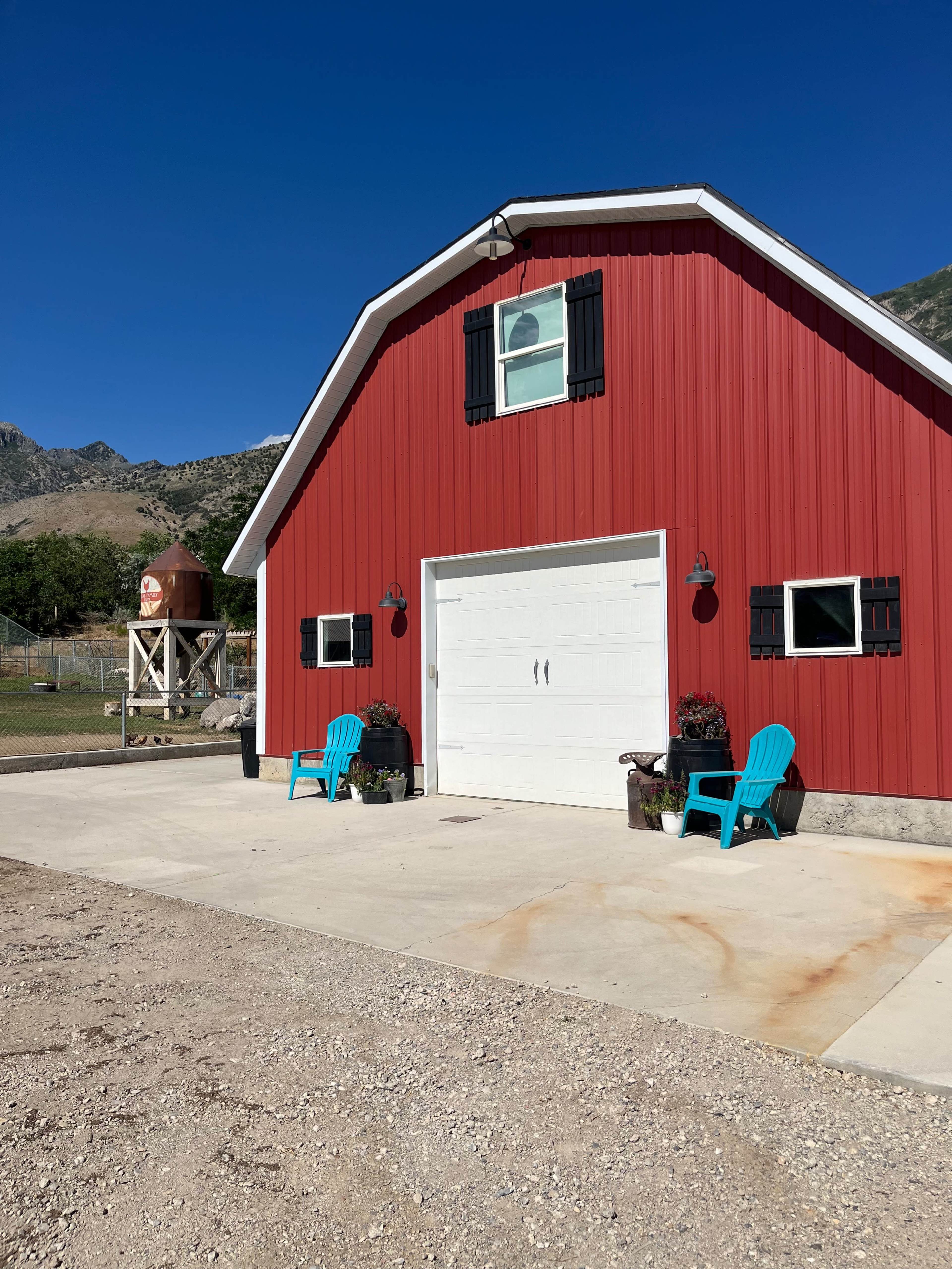 A red barn with a white garage door, two turquoise chairs in front, and a water tank nearby under a clear blue sky.