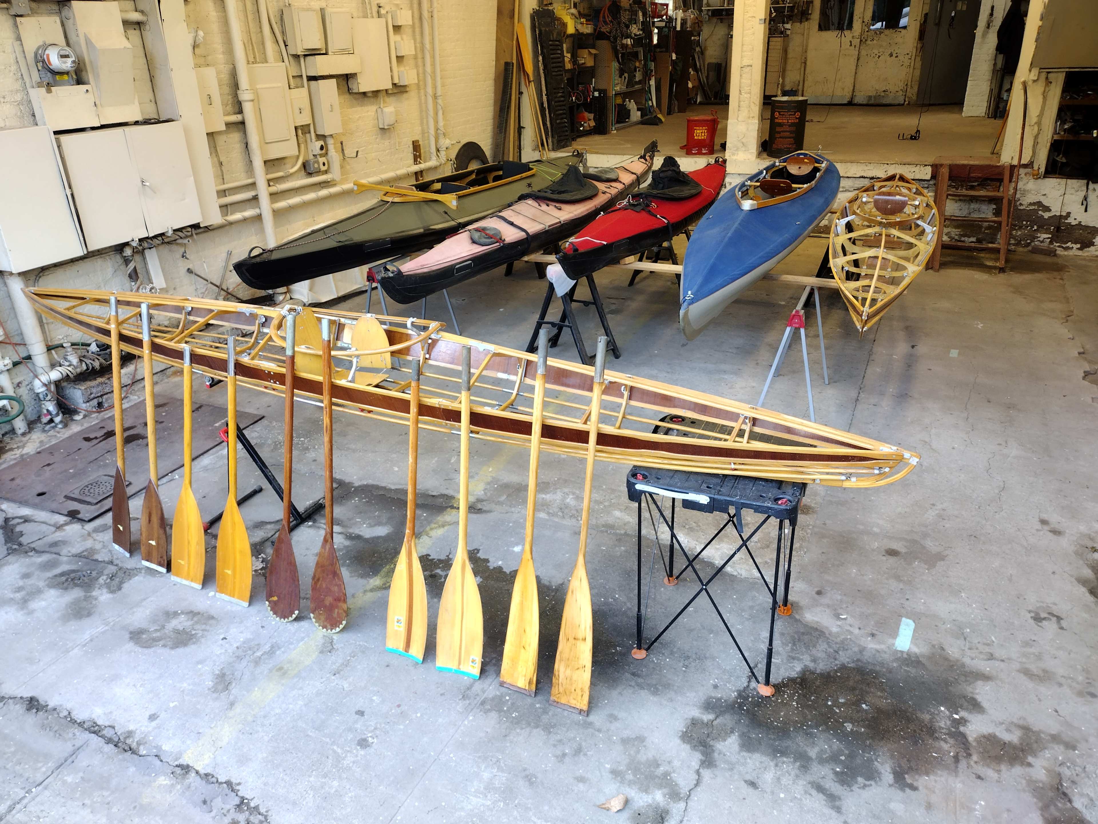 A set of kayaks and a wooden kayak frame with paddles are displayed in a spacious workshop.