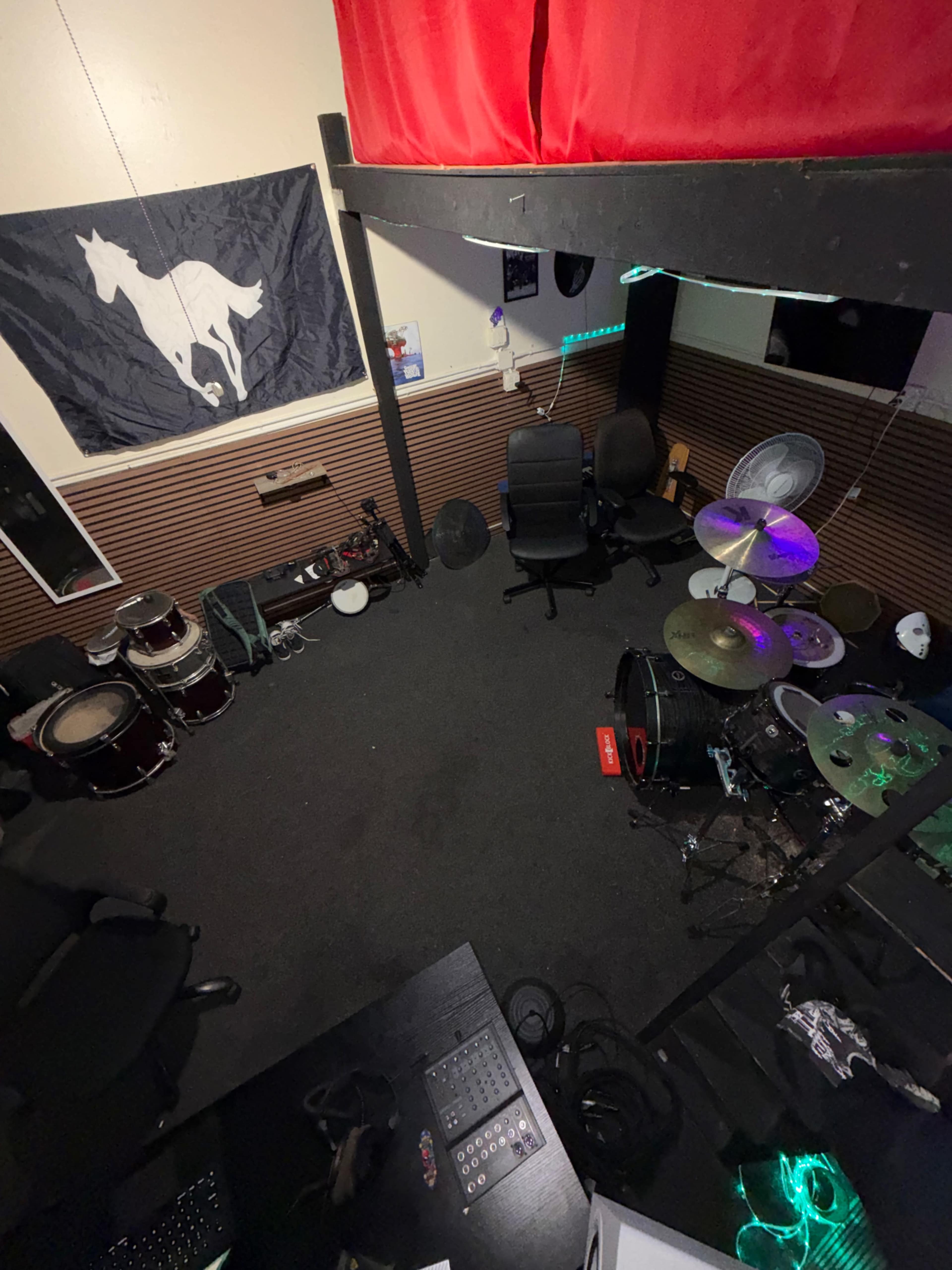 The image shows a music rehearsal space featuring a drum set, several chairs, and a wall decorated with a flag depicting a horse.