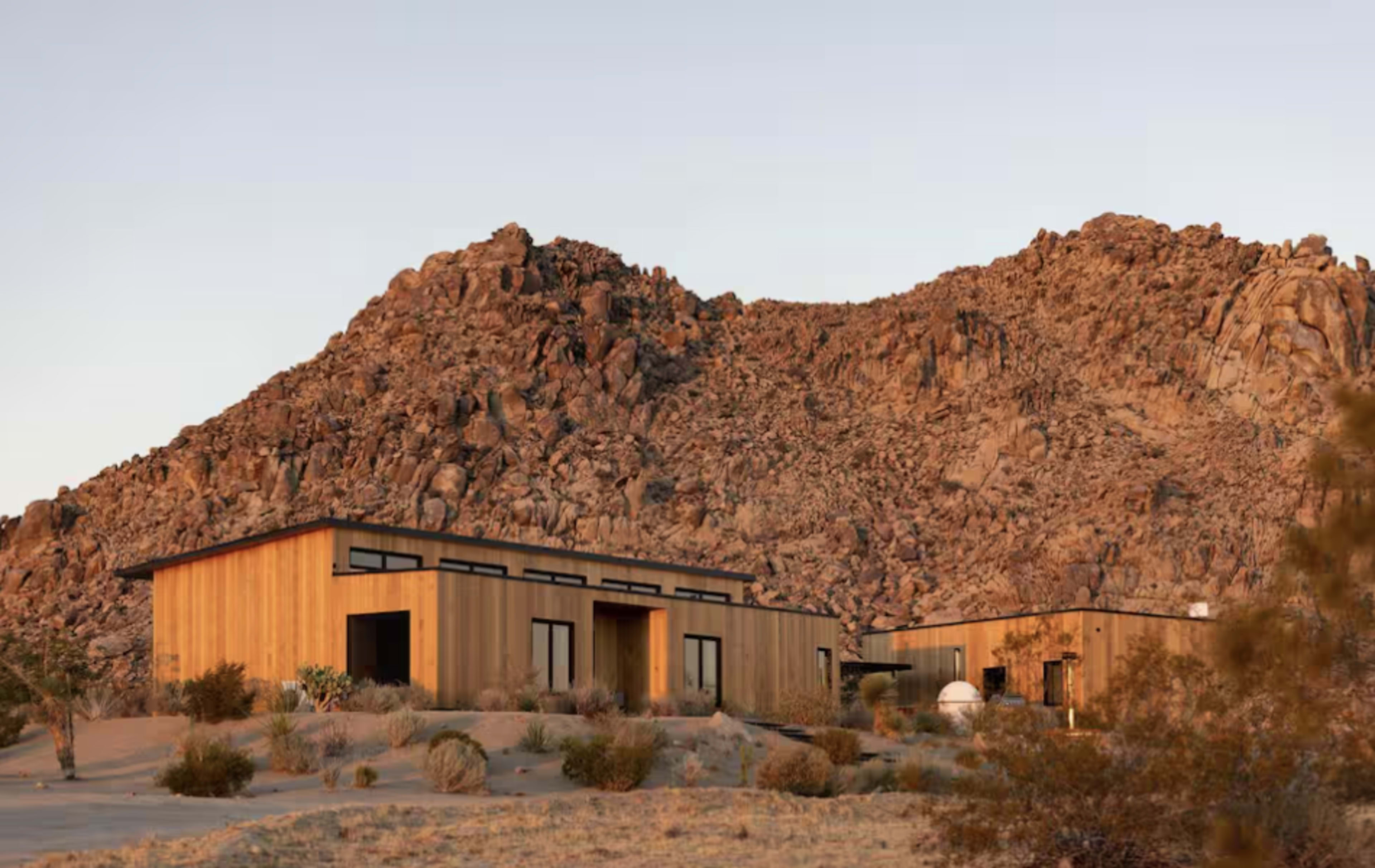 Two modern wooden houses are situated in a desert landscape near a rocky hillside.