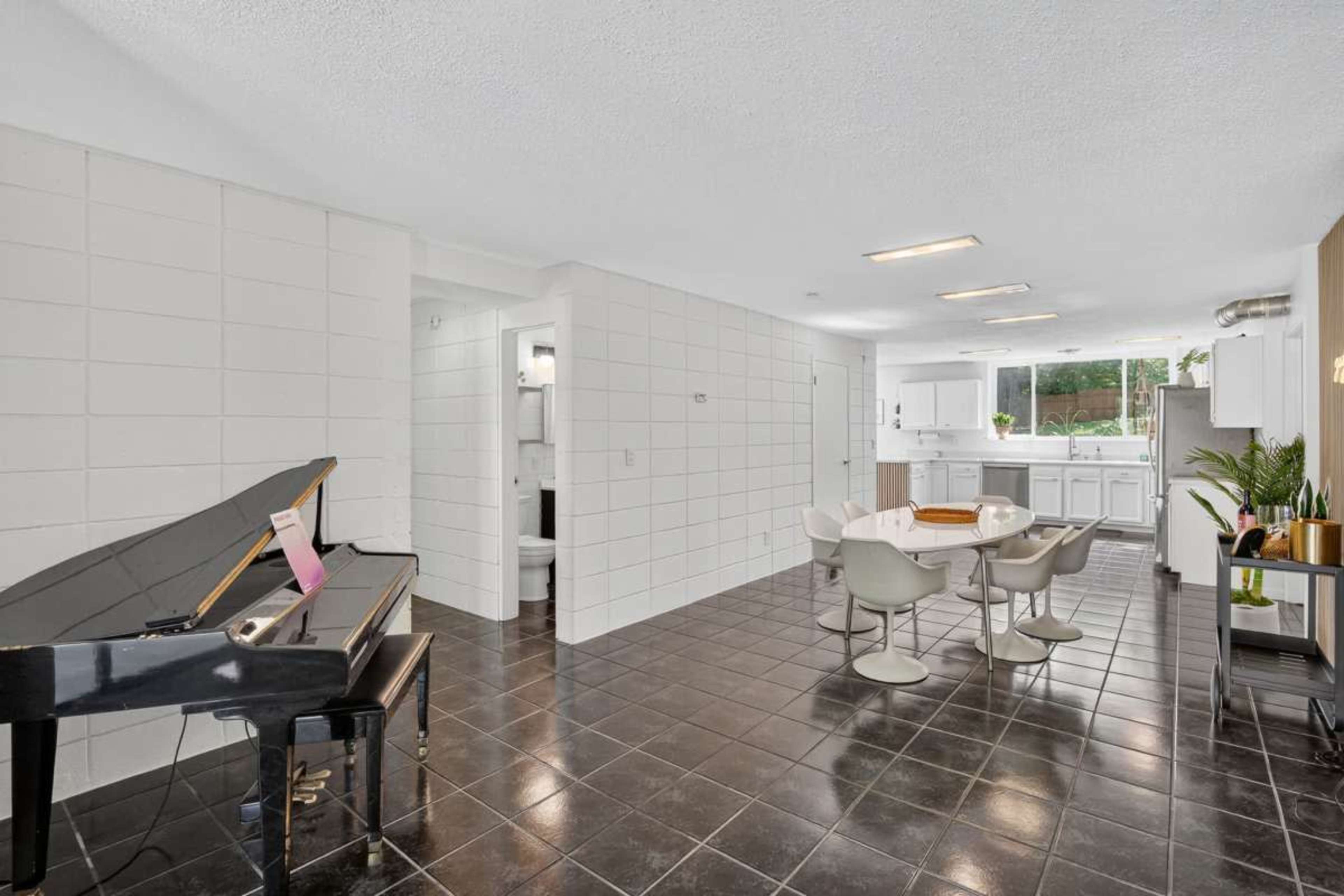 The image showcases a modern, open-concept space featuring a black piano, a dining table with chairs, and a view of a kitchen with white cabinetry.