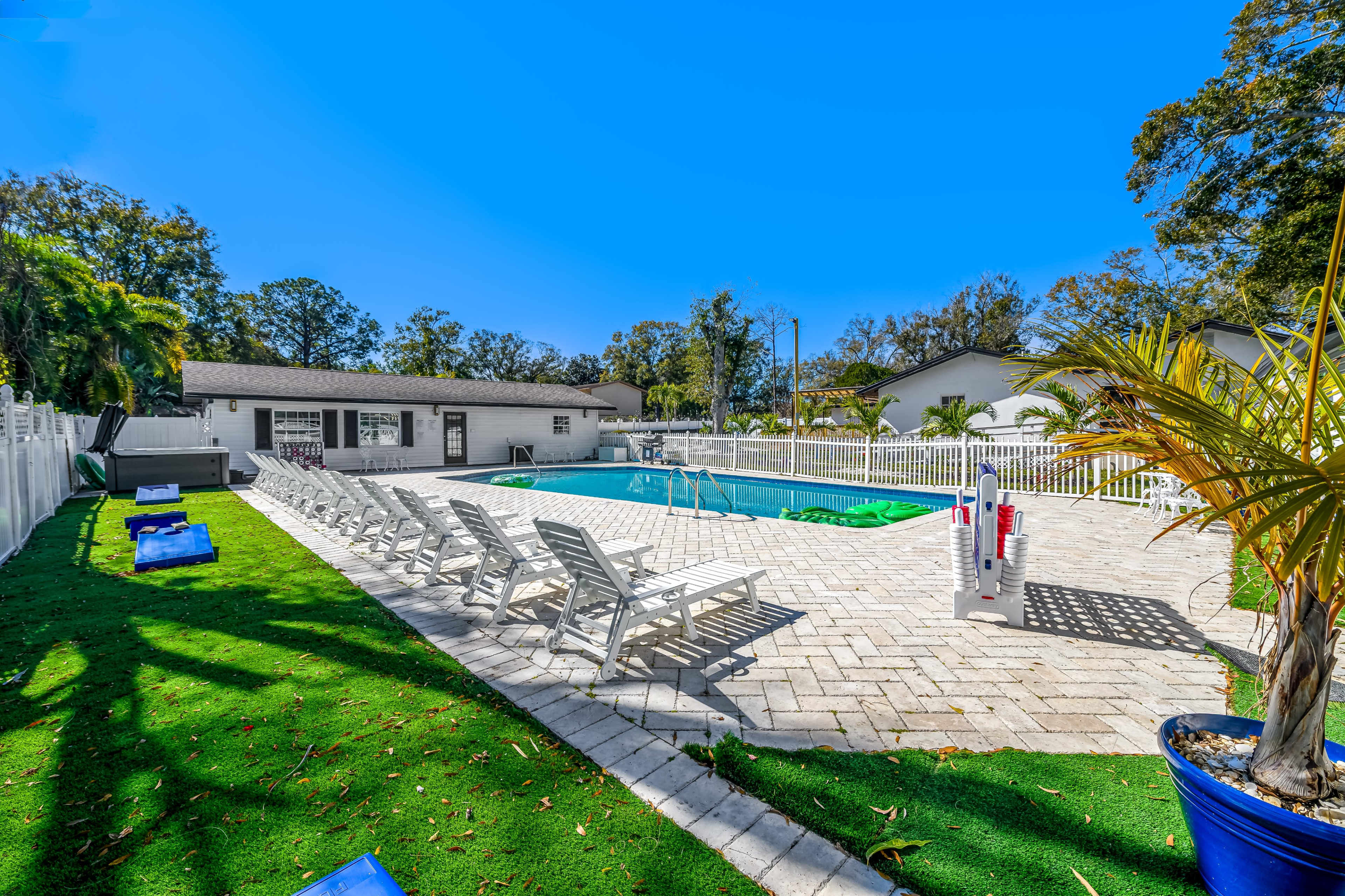 The image shows a swimming pool area with lounge chairs and a well-maintained patio surrounded by greenery and a white fence.