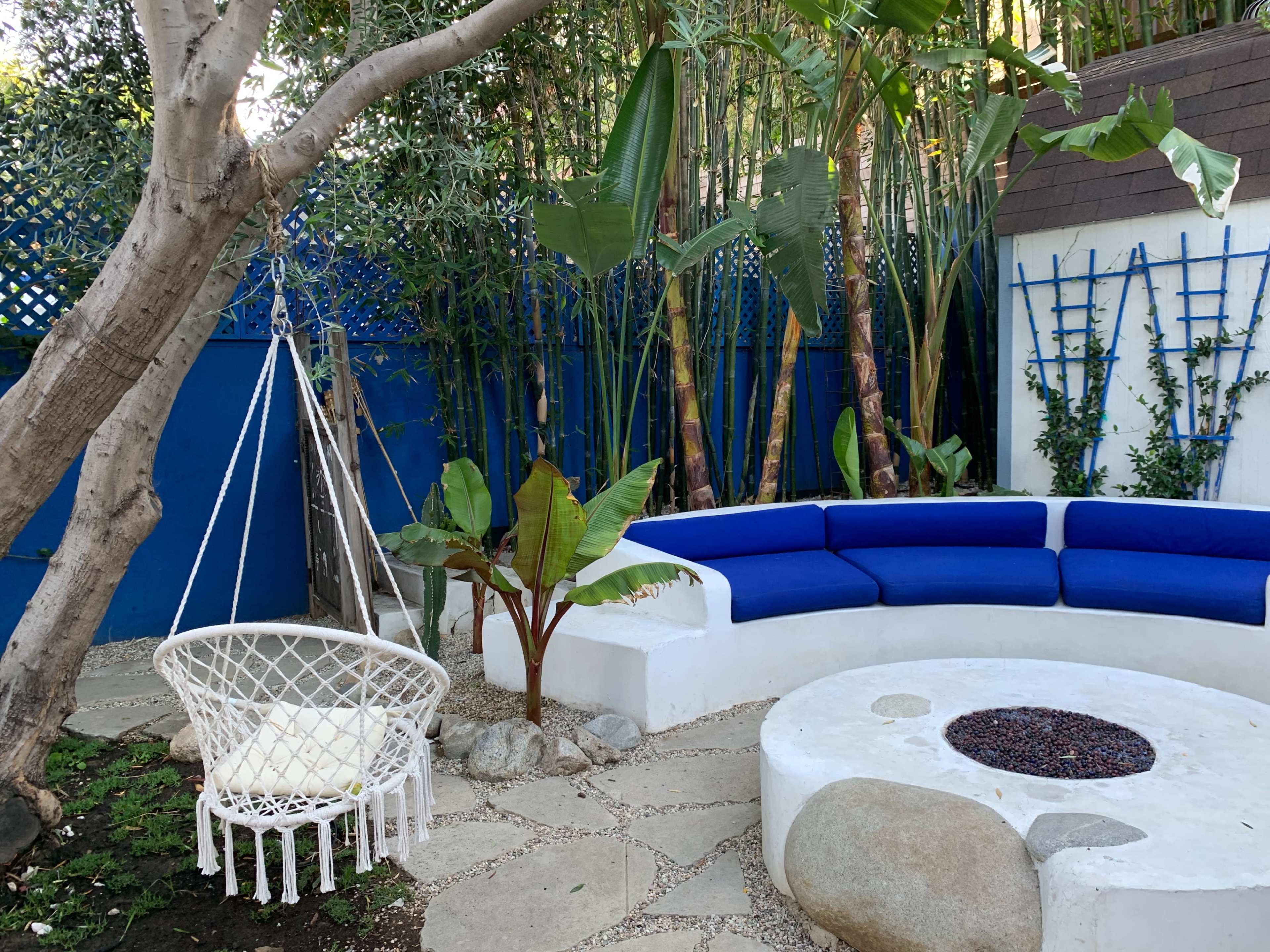 The image shows a cozy outdoor space featuring a circular stone fire pit, a semicircular white seating area with blue cushions, and a hanging wicker chair surrounded by lush greenery.