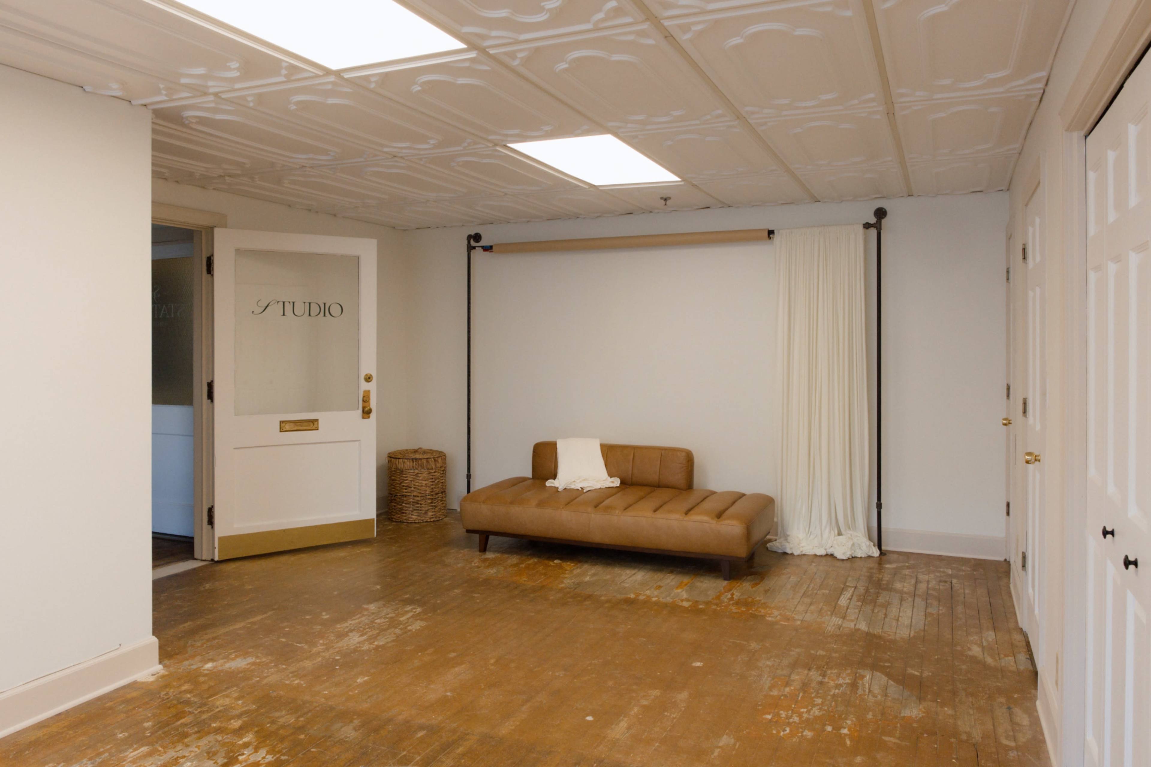The image shows a spacious room with a wooden floor, a brown couch, a white curtain on a rod, and a door labeled "STUDIO."