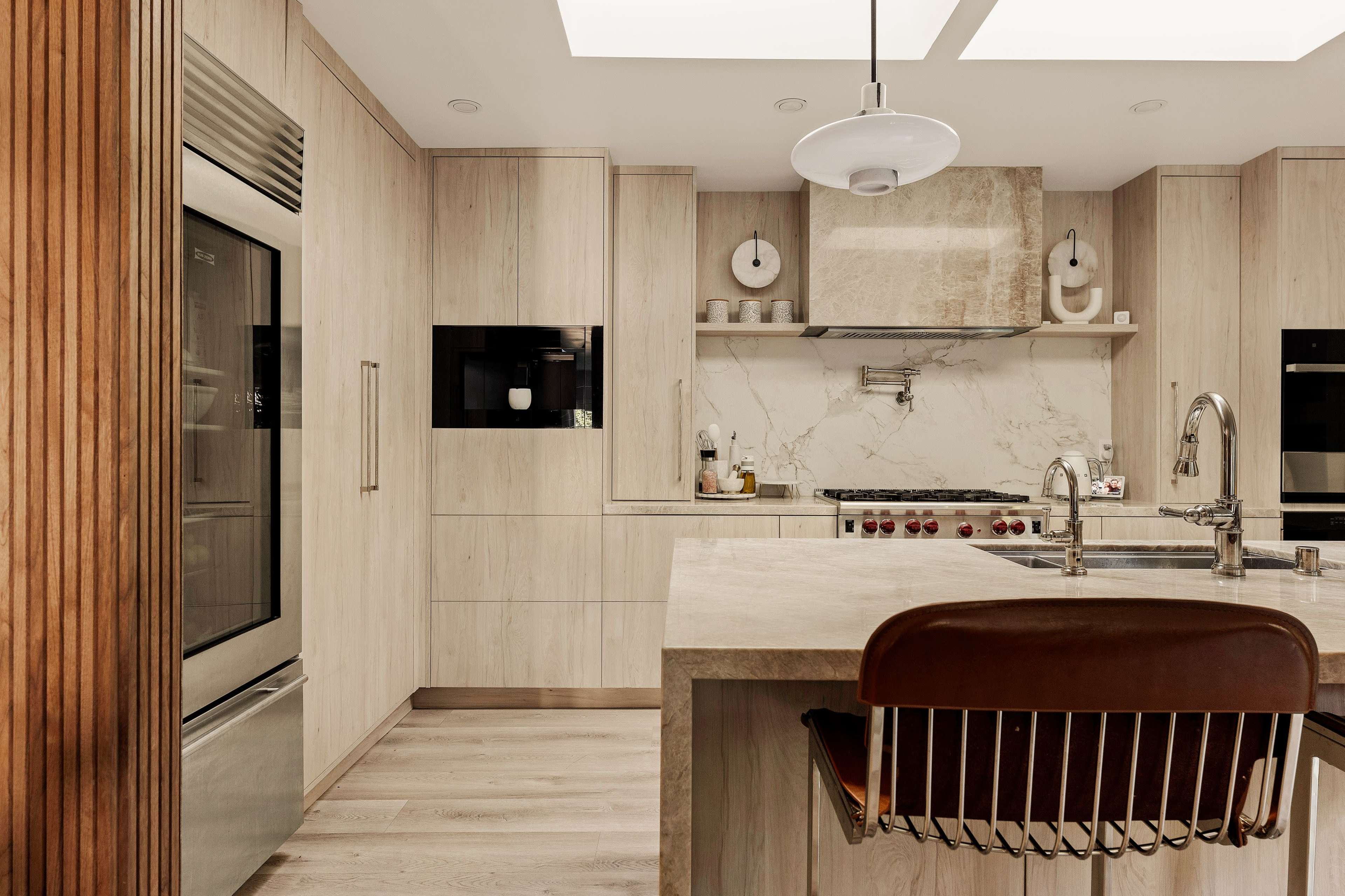 The kitchen features light wood cabinetry, a large island with a stone countertop, stainless steel appliances, and a central pendant light.