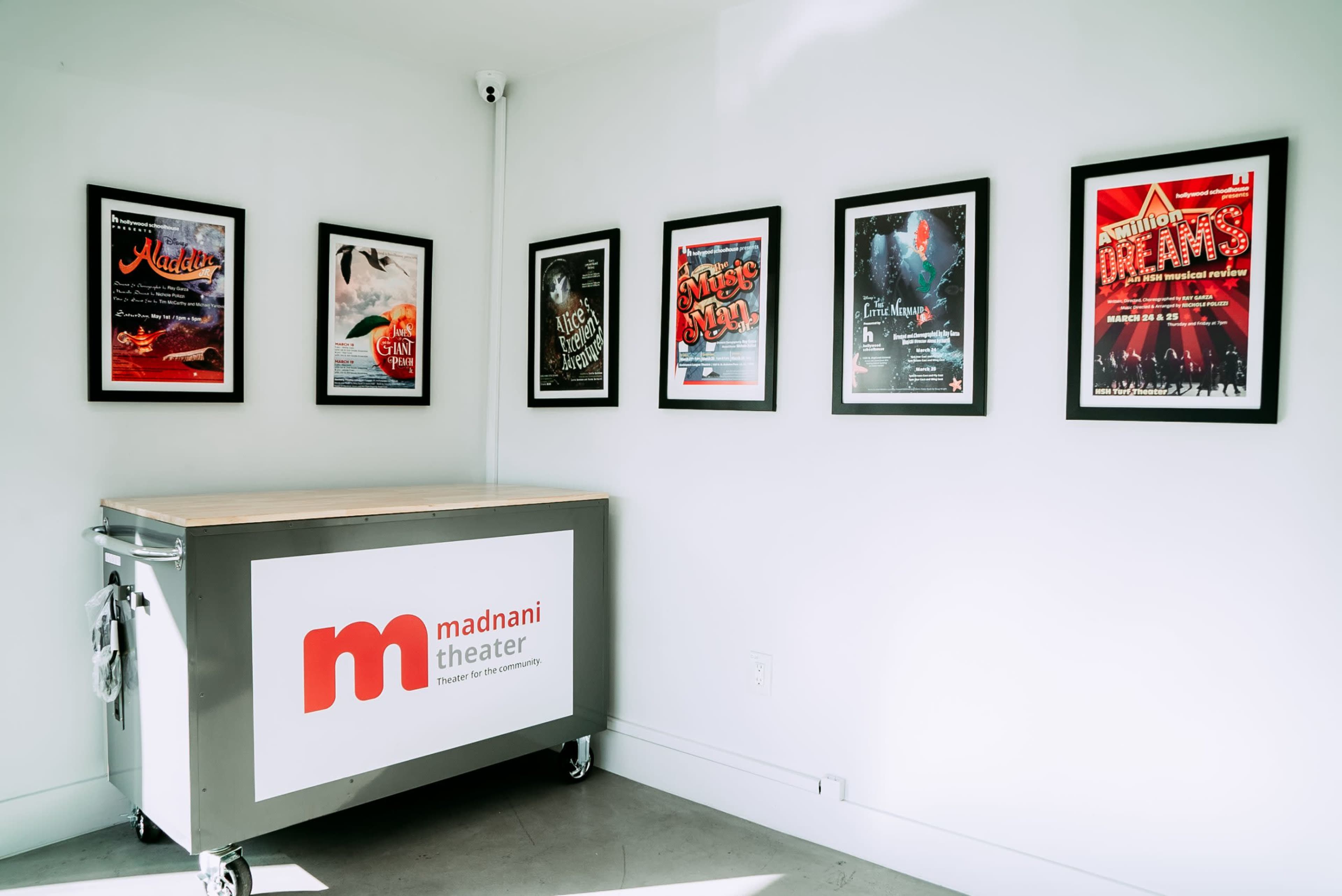 The image shows a white-walled room featuring a series of framed posters on the wall and a storage cart labeled "madnani theater" in front.