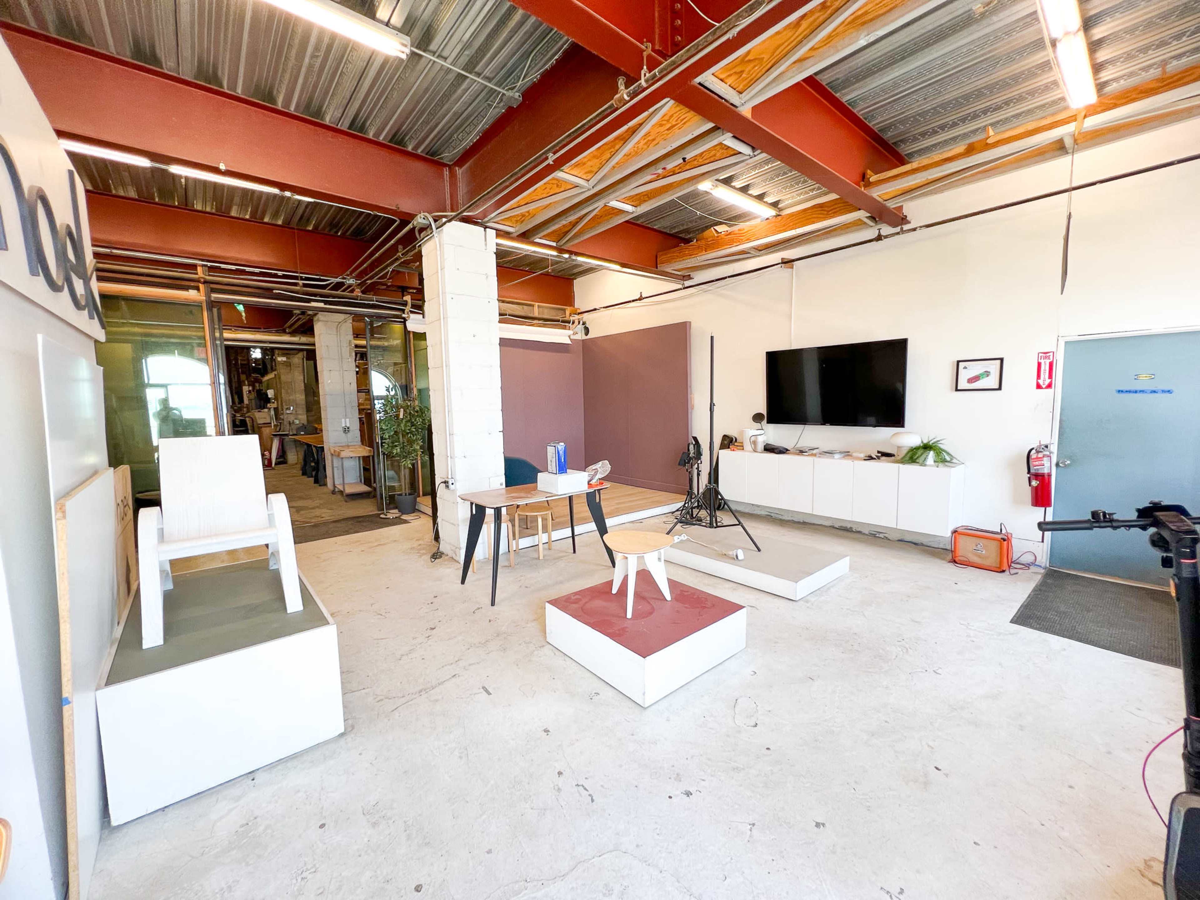 The image shows an industrial-style interior space with a minimalist design featuring furniture, a television mounted on the wall, and a large entrance.