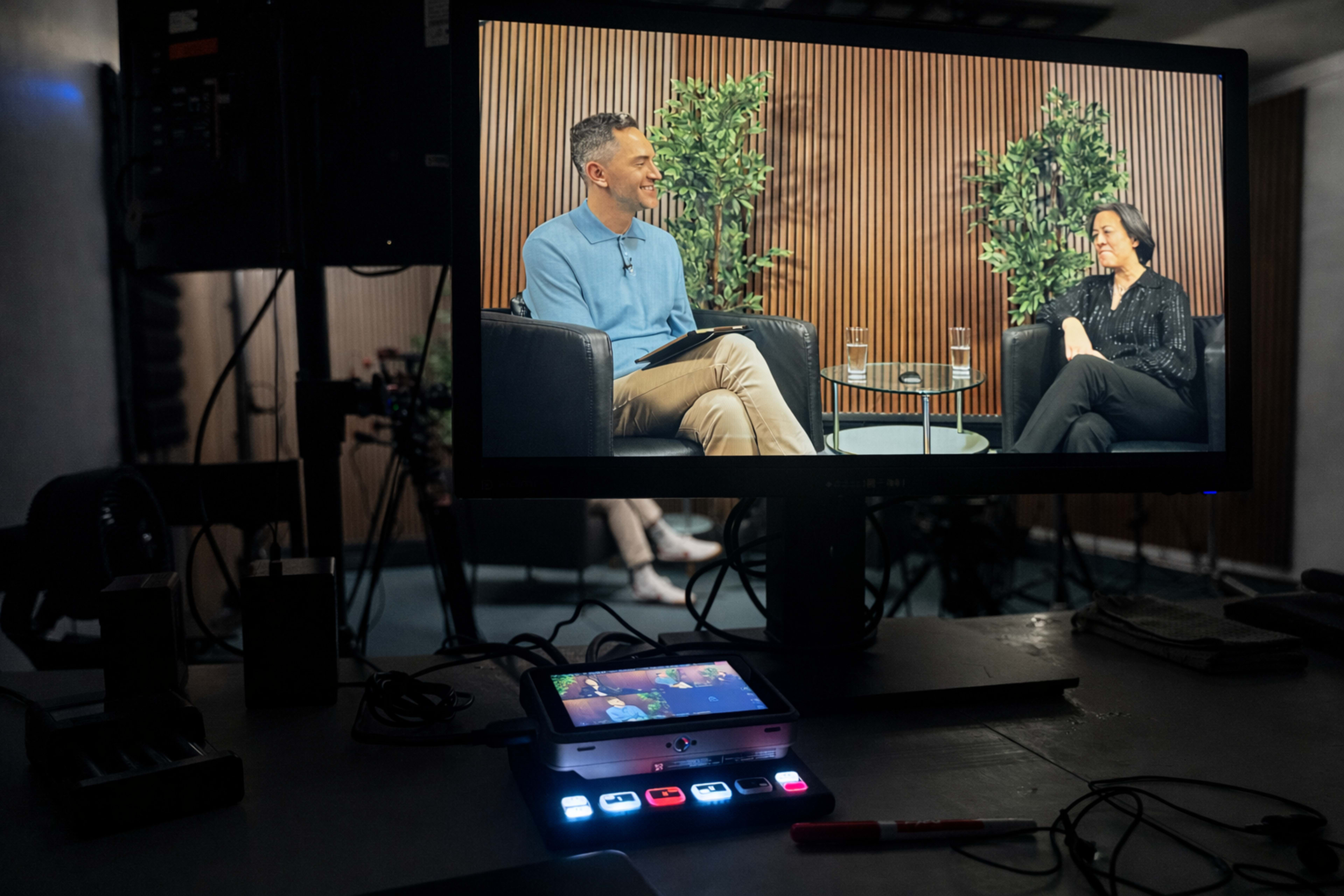 A television screen displays two individuals engaged in an interview, while a control panel with buttons is positioned in the foreground.