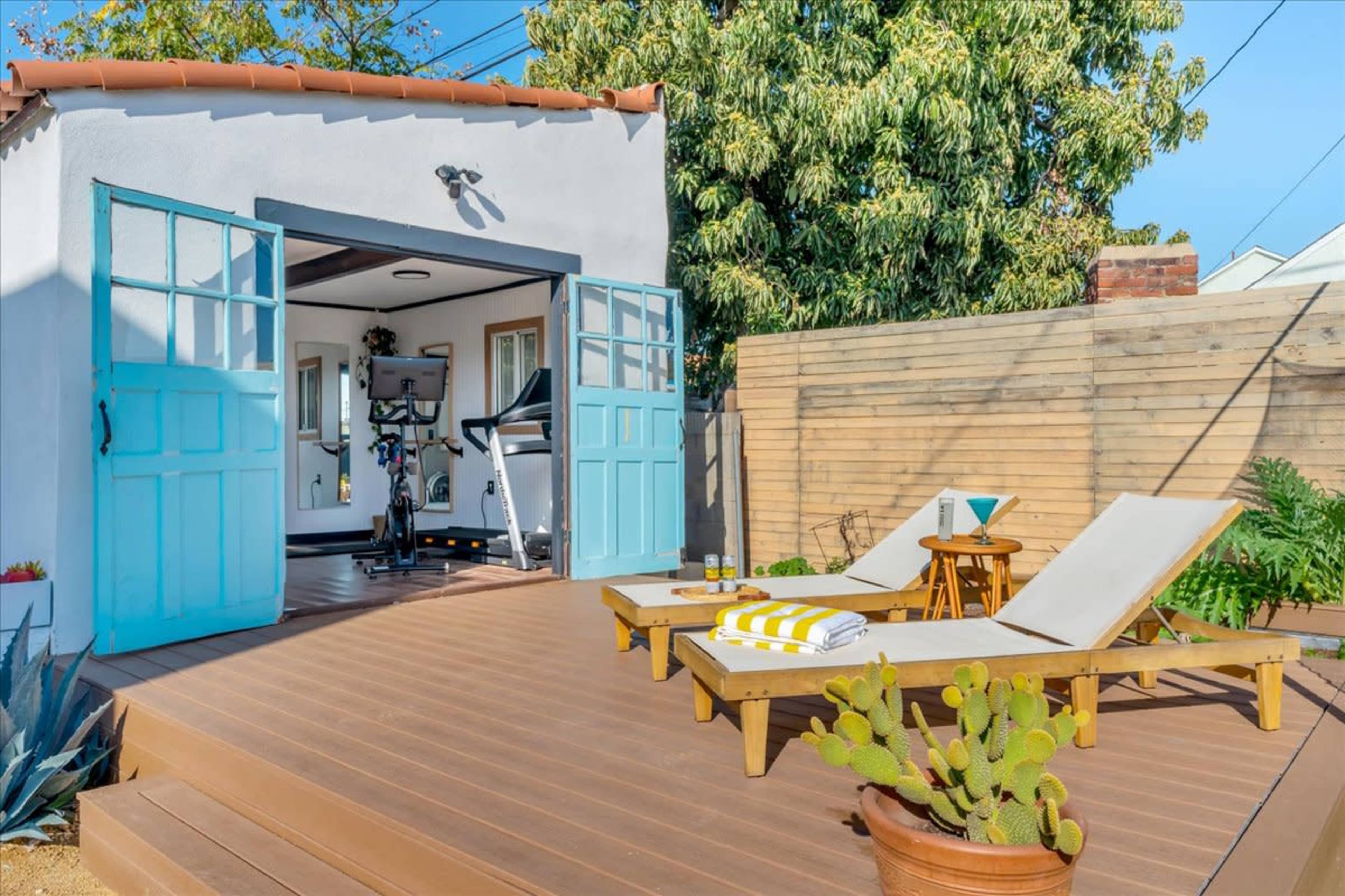 A sunny outdoor deck features two lounge chairs, a cactus in a pot, and a small table with yellow-striped items, leading to a room with open blue doors that contains exercise equipment.