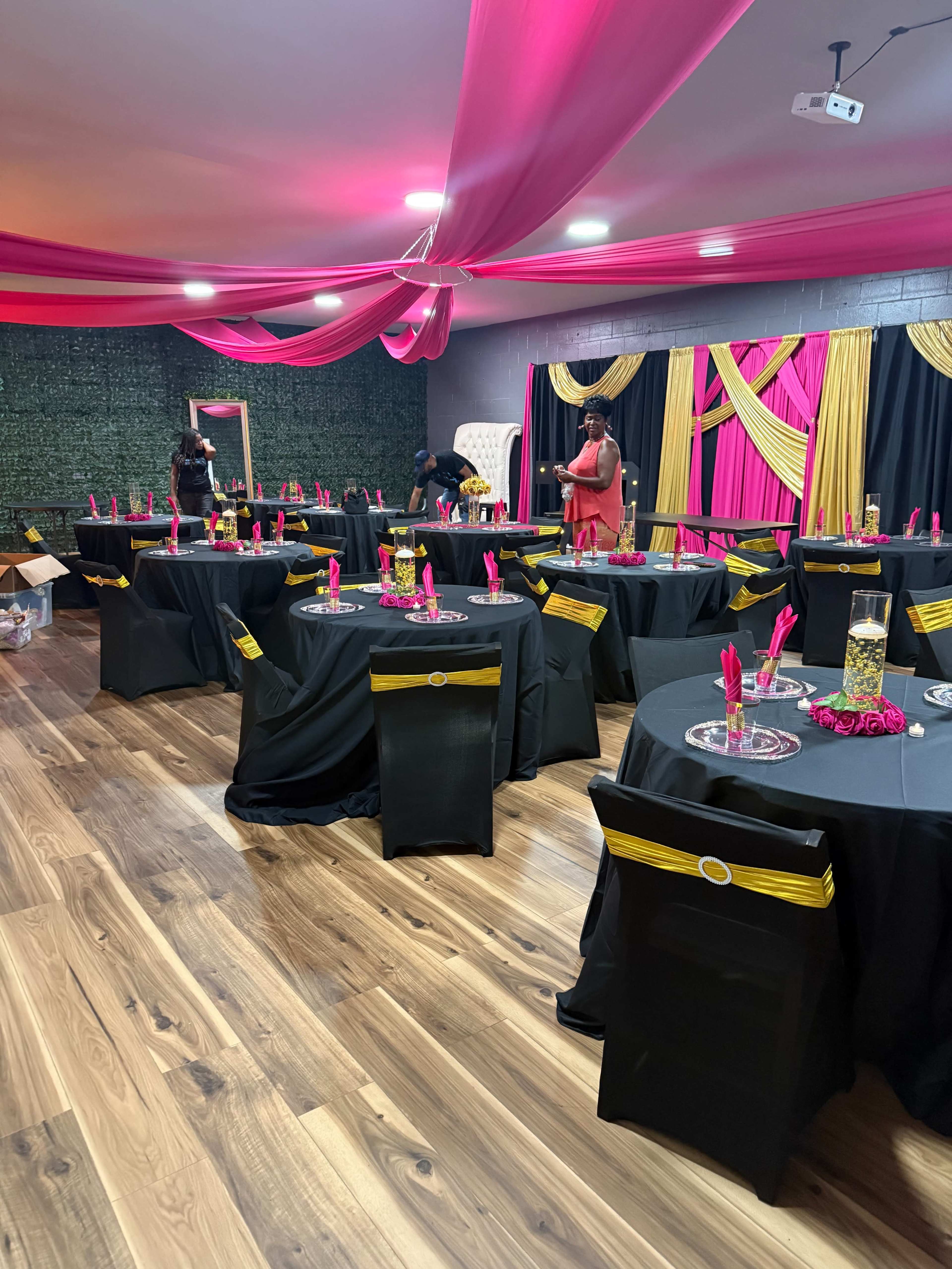 The image shows a decorated event space with black tables covered in black tablecloths and adorned with pink napkins, colorful centerpieces, and vibrant drapes on the walls.