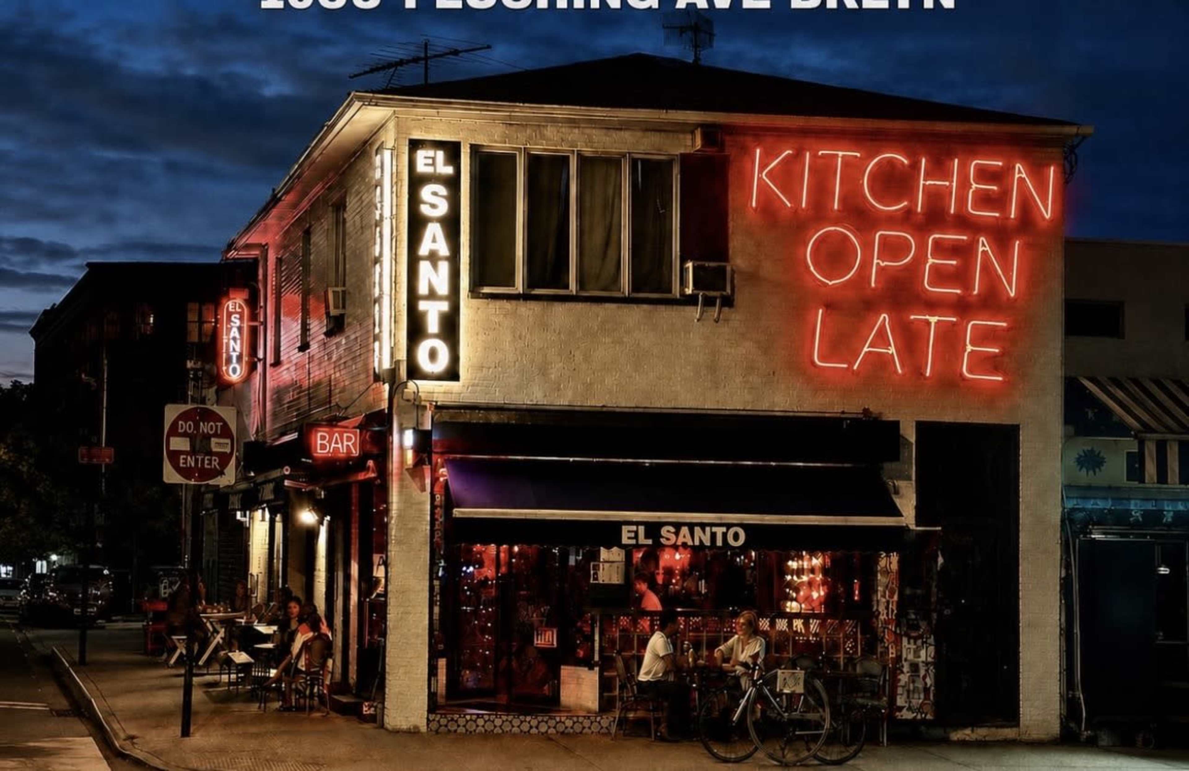 The image shows a brightly lit restaurant and bar called "El Santo" on a street corner in Brooklyn, featuring neon signs that read "KITCHEN OPEN LATE."