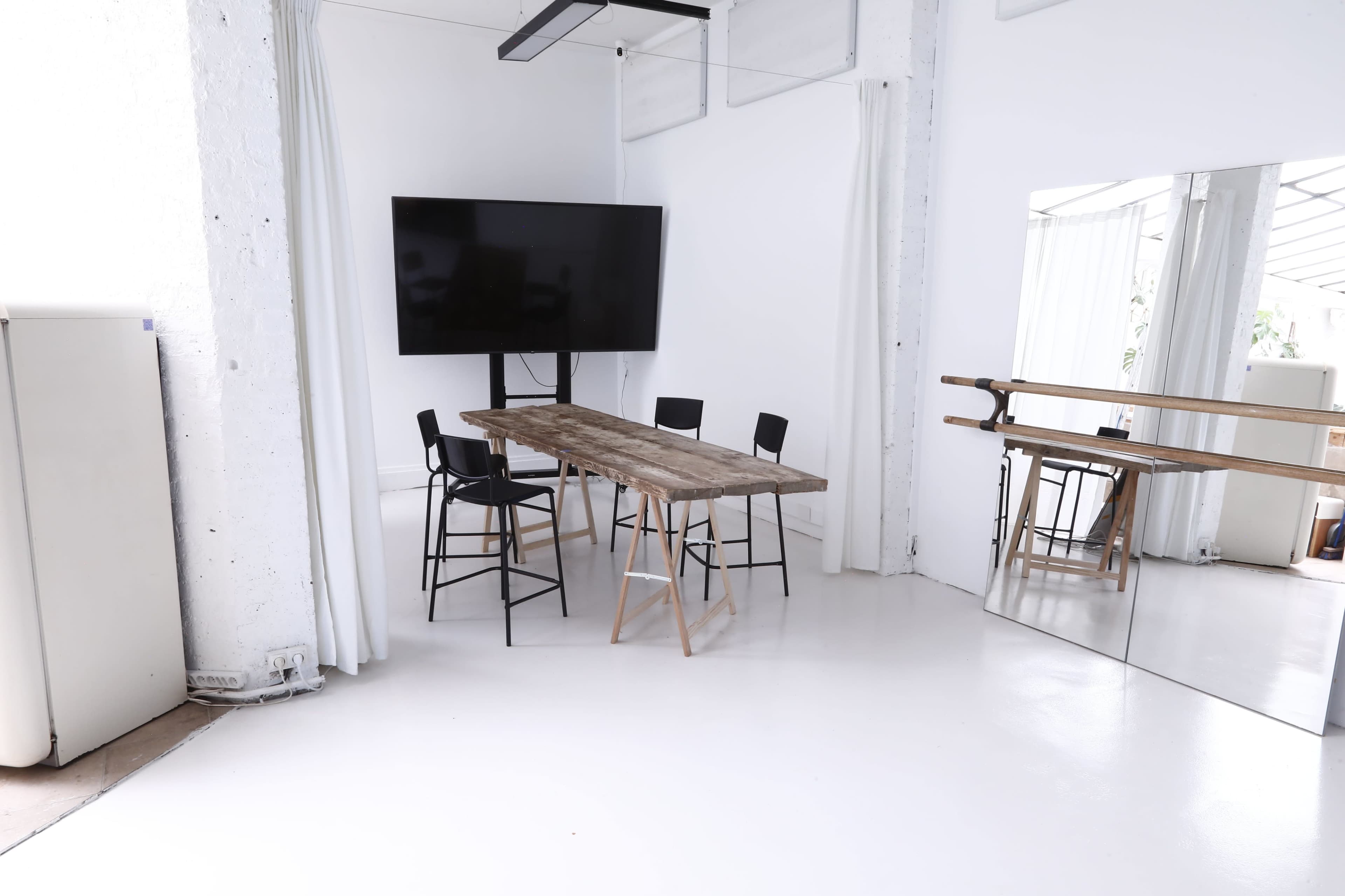 A modern meeting room features a wooden table surrounded by black chairs, a large television on the wall, and a mirrored surface in the corner.