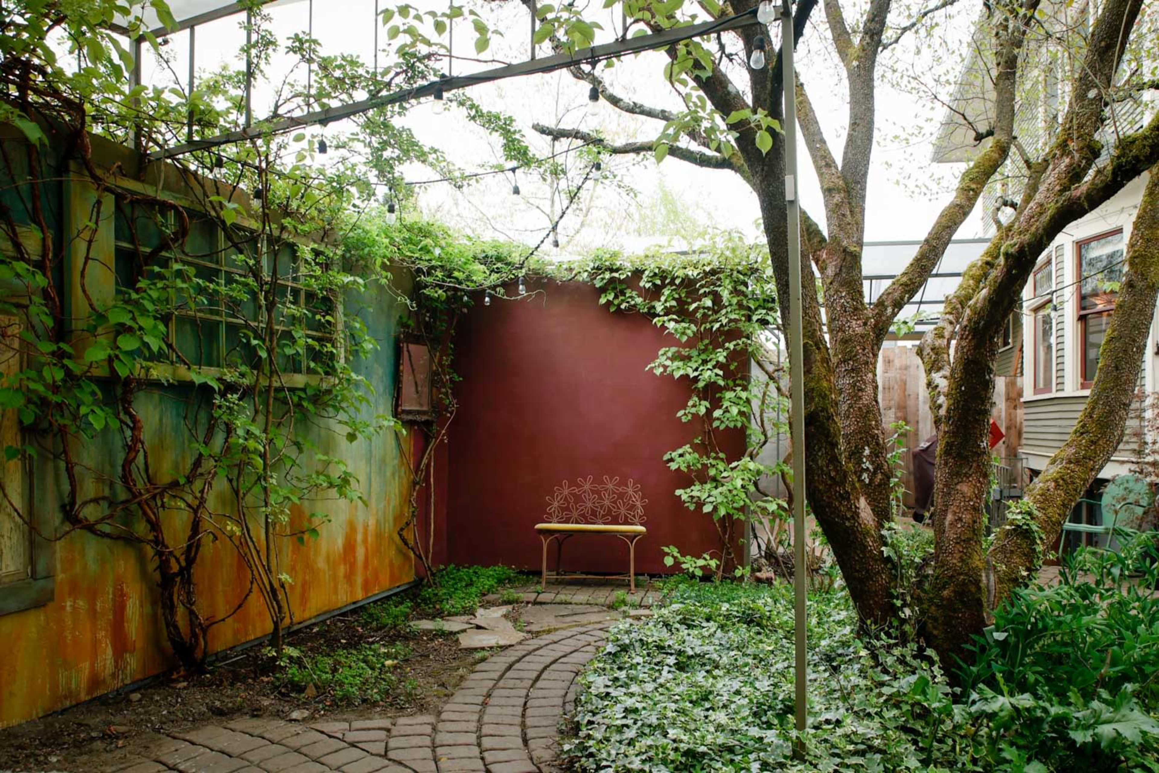 A garden space with a curved stone path leading to a metal bench surrounded by greenery and a vine-covered wall.